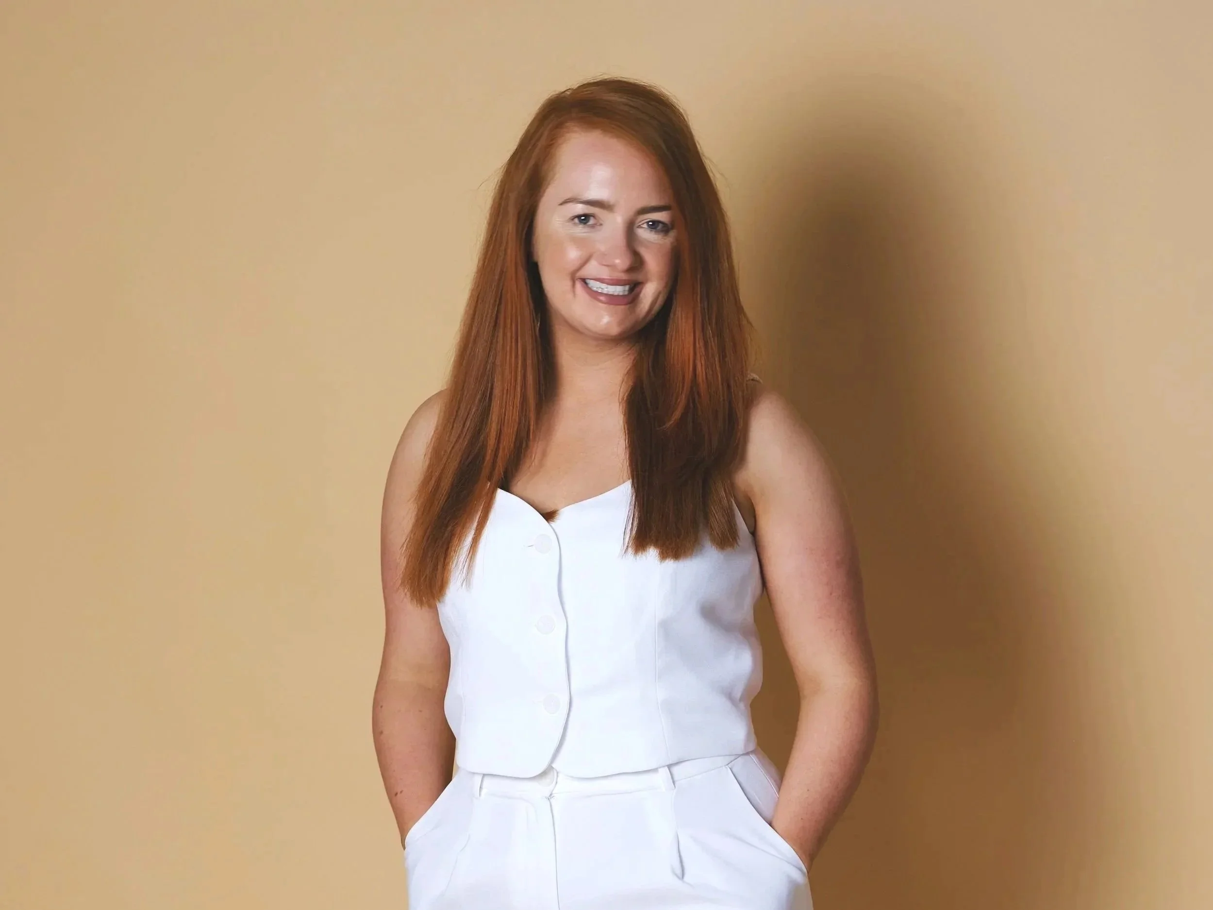 Photo of Sound Waves Founder, Dee O'Kelly, wearing a white sleeveless top and white high-waisted pants, standing against a beige background, smiling with hands in pockets, wearing black and white sneakers.