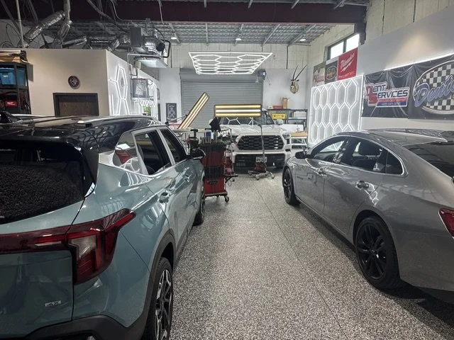 Inside an auto repair shop with two silver cars parked inside, one SUV on the left and one sedan on the right. The background shows various tools, equipment, and a partially open garage door, with bright lighting and a modern design interior.