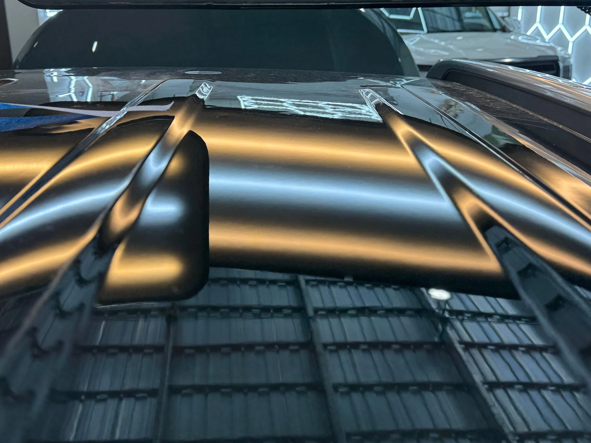 Close-up view of a vehicle's hood with reflections, showing parts of the windshield and other cars in the background, in a showroom with neon lighting.