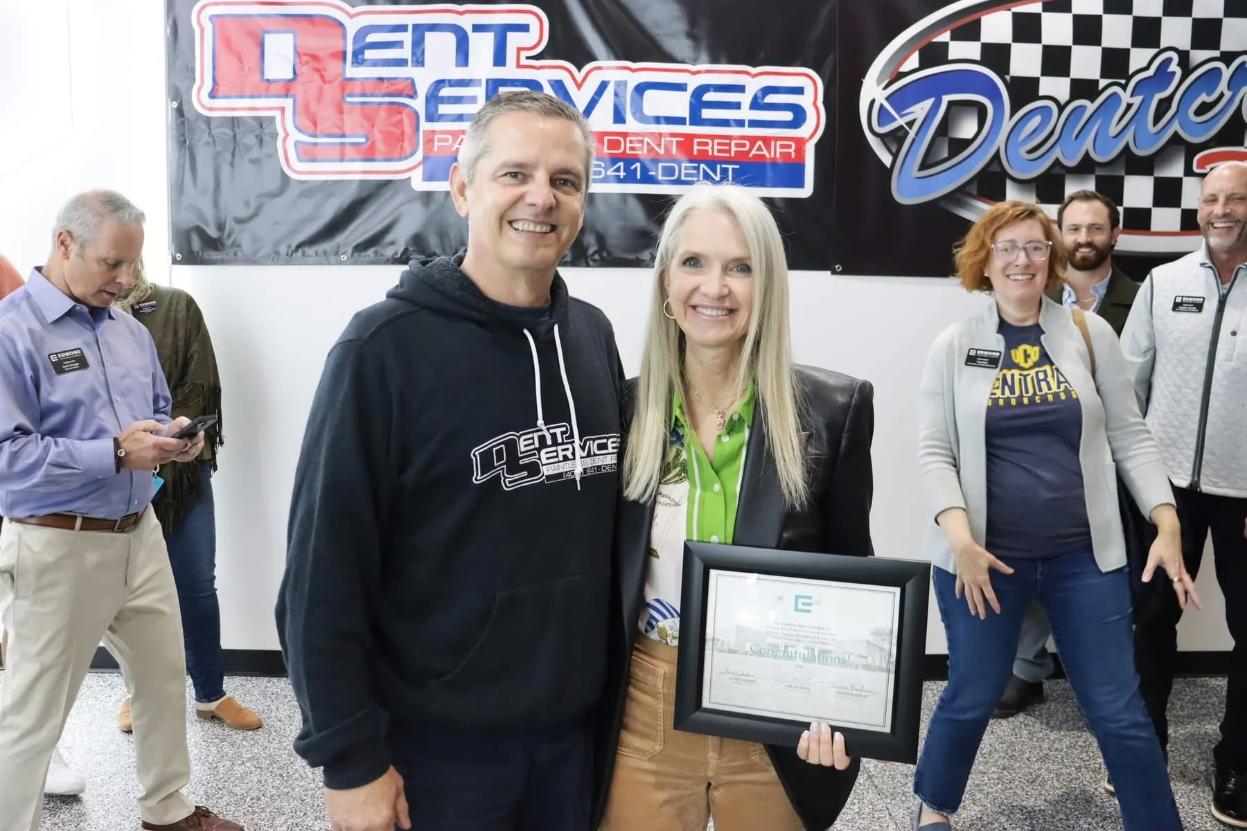 A group of people at an event, with a man and a woman in the foreground holding a framed certificate and smiling. Behind them, banners display business names, including Dent Services, and other people are visible in the background.