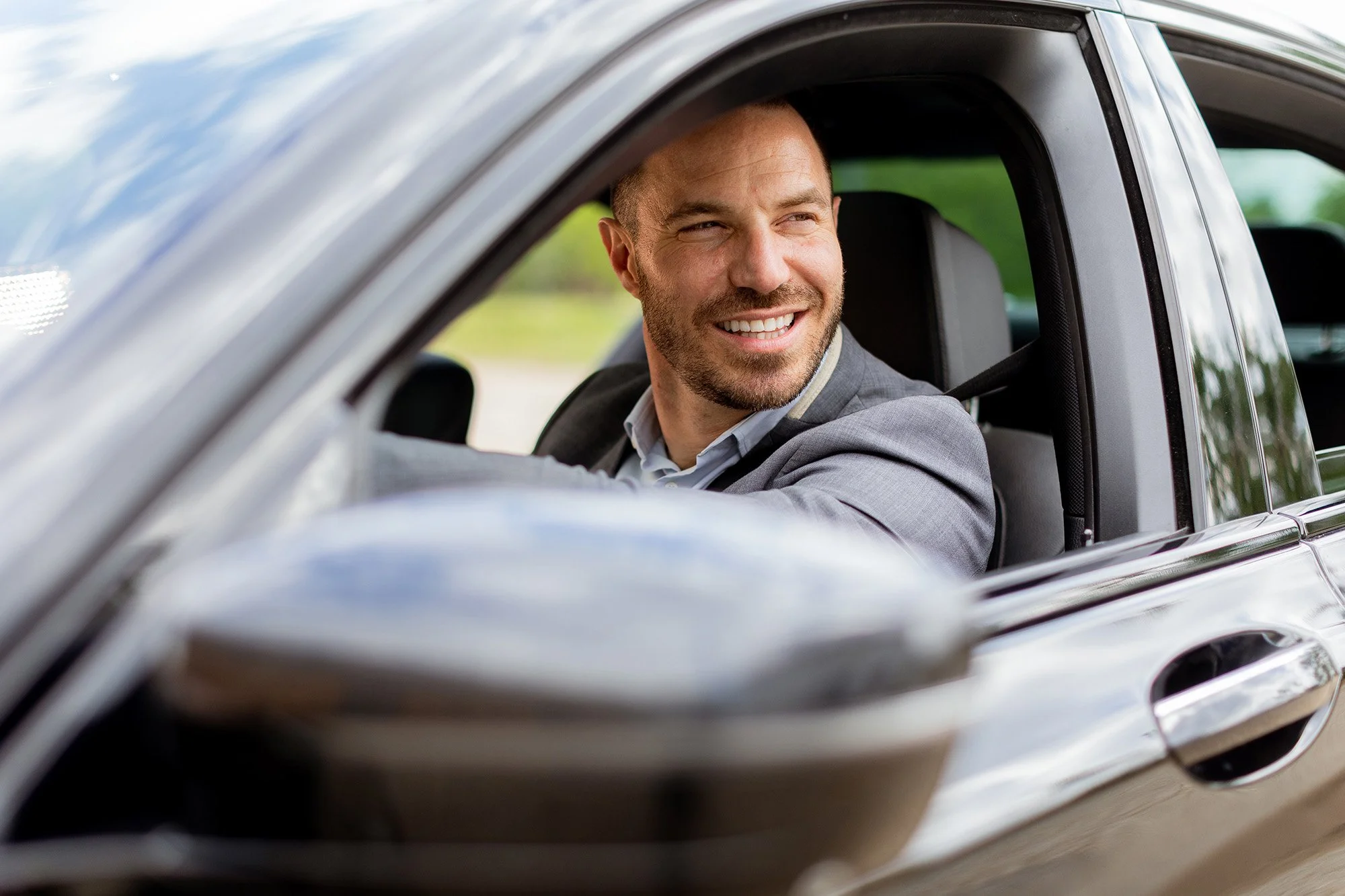 A man with a beard and short hair smiling while sitting in the driver’s seat of a silver car, looking out the window.