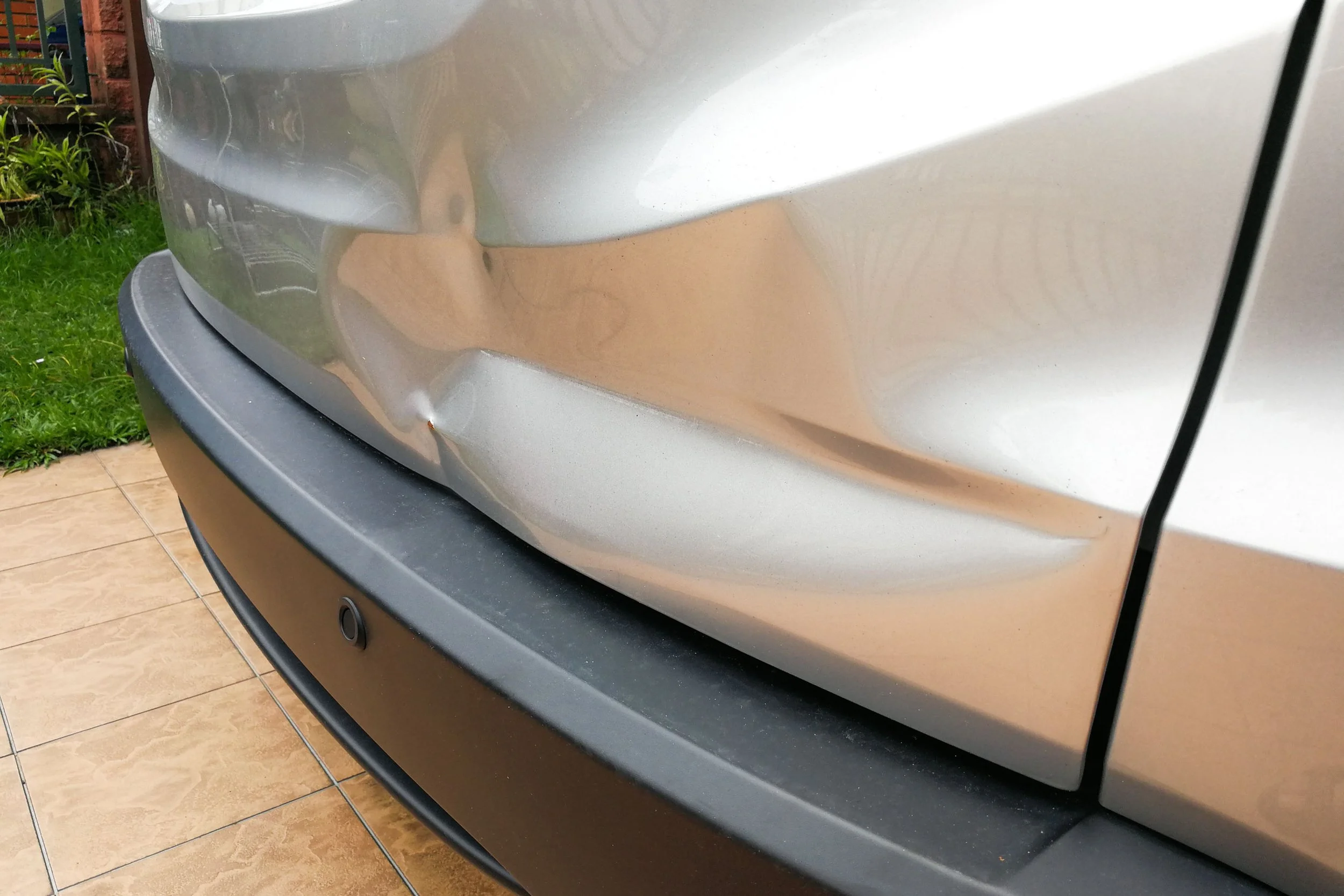 Close-up photo of the rear corner of a silver car, showing a slight dent, part of the taillight, bumper, and a parking sensor on the black lower bumper.
