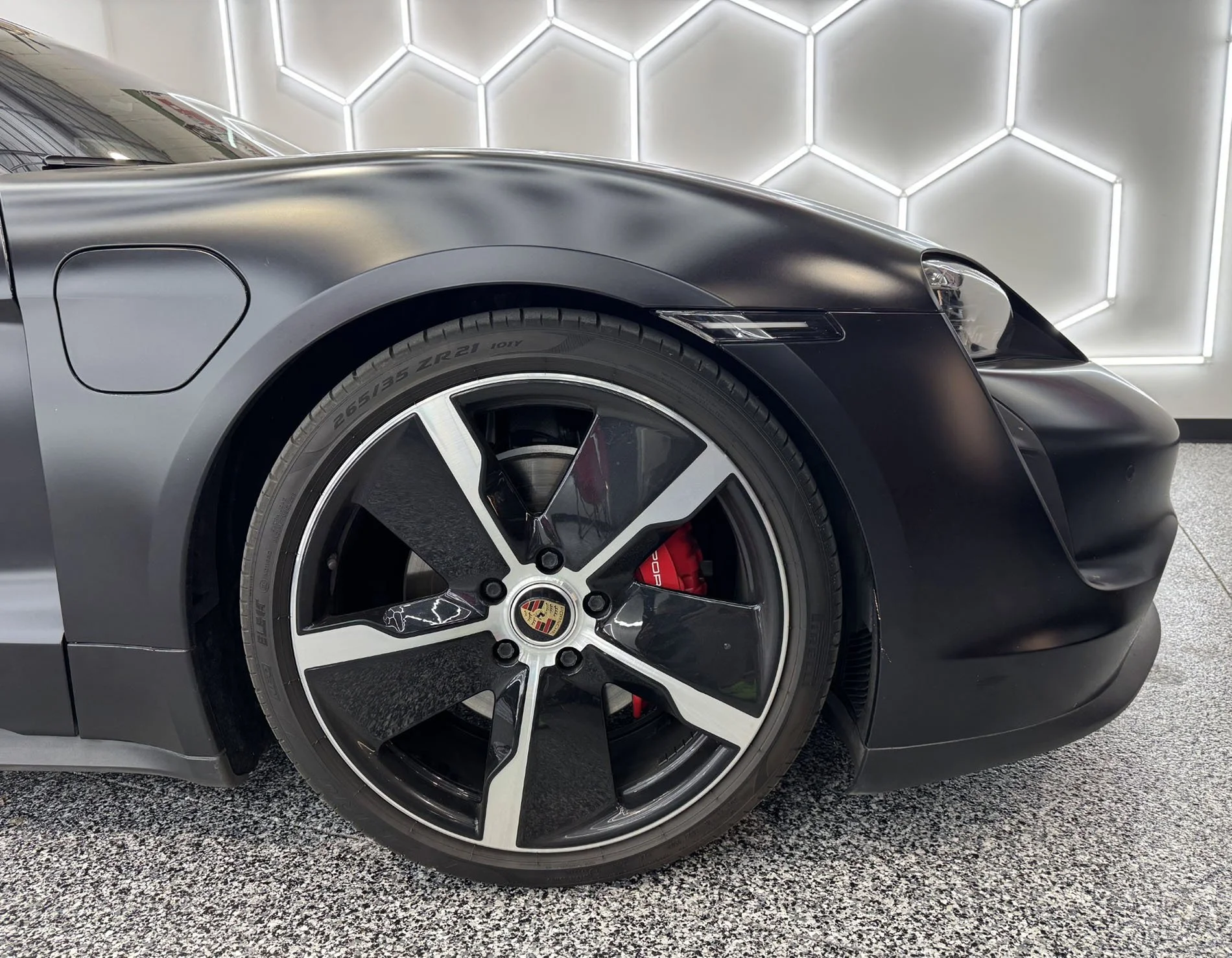 Close-up of a black sports car's front left side, including the wheel with a Porsche emblem, and part of the car's sleek body with a hexagonal patterned illuminated wall in the background.