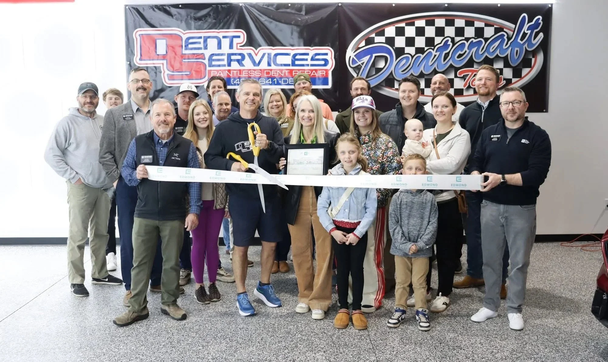 A group of people at a grand opening event, holding large ceremonial scissors with a ribbon in front of them. There are adults and children smiling for the camera, with banners advertising in the background.