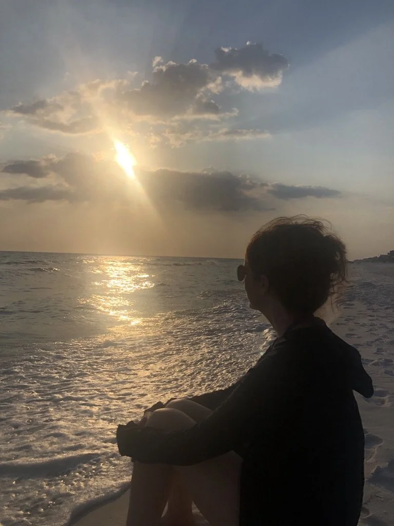 Person sitting on the beach during sunset, looking at the ocean with the sun partially covered by clouds.