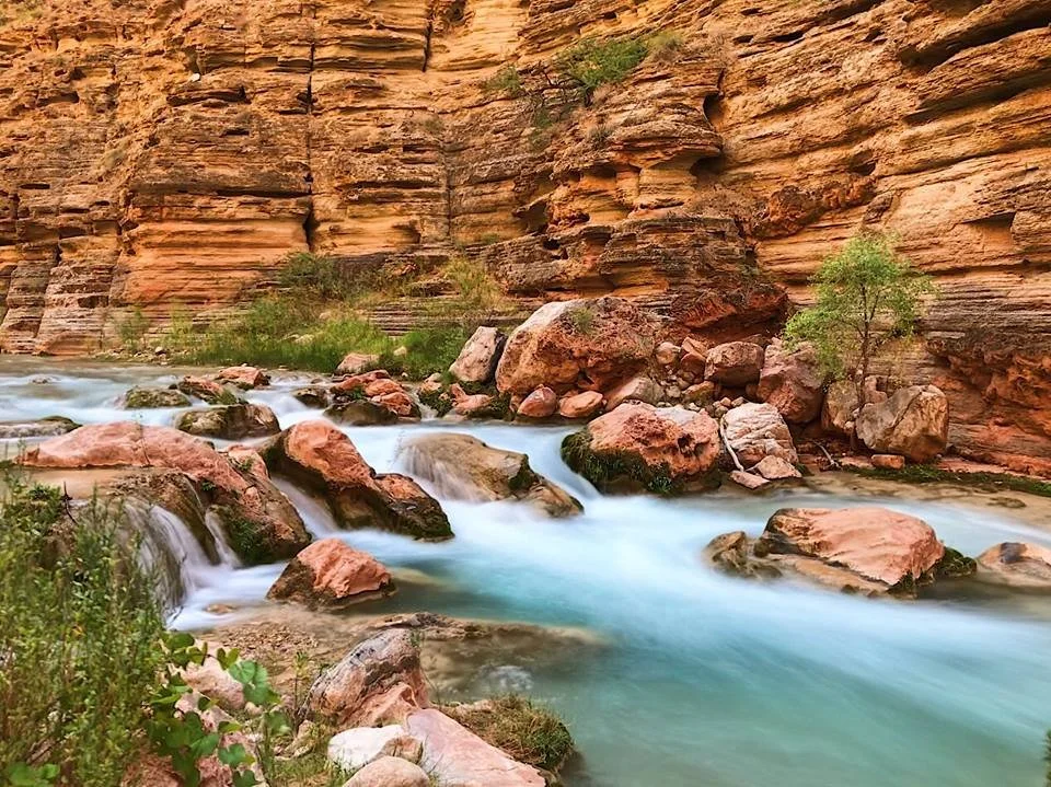 Rocks and small waterfalls in a river with steep, reddish rock cliffs and sparse green vegetation.