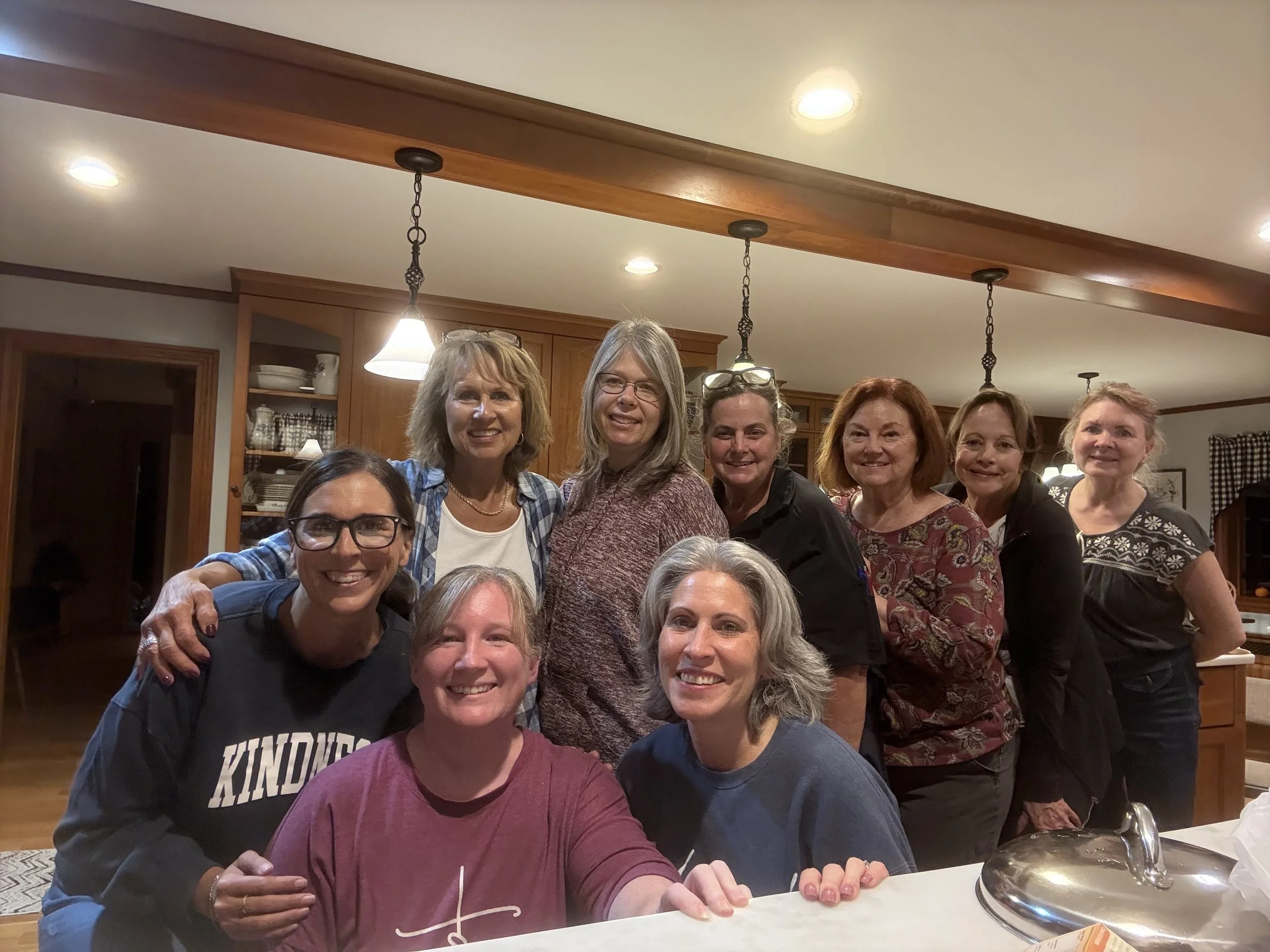 A group of women gathered around a table inside a home, smiling and posing for a photo. The setting appears to be a kitchen or dining area with wooden cabinets and hanging lights.