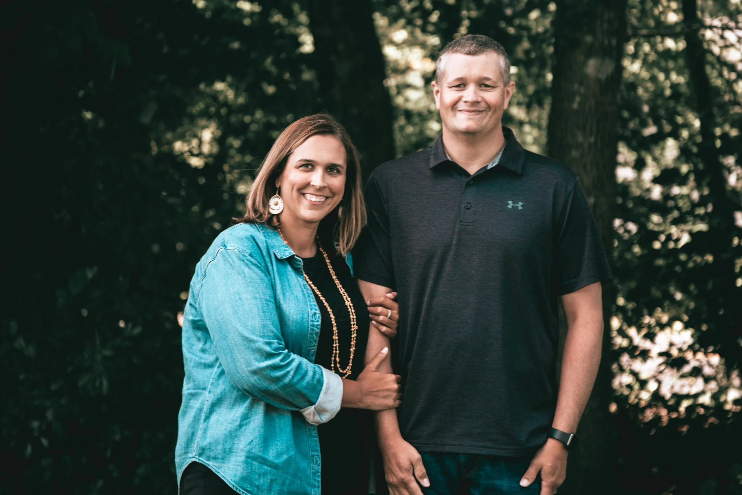 A woman and a man smiling outdoors surrounded by trees, with the woman holding the man's arm.