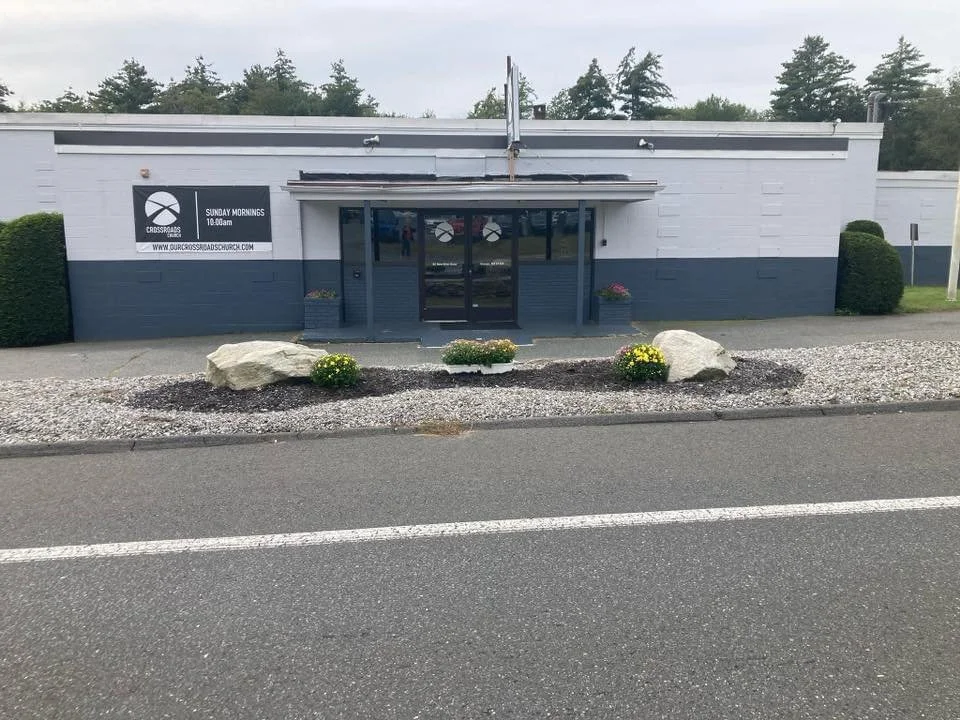 Front of a church with a sign that reads "Crossroads Church" and indicates Sunday service at 10:00 AM, with a glass entrance, rock landscaping, and hedges on either side.