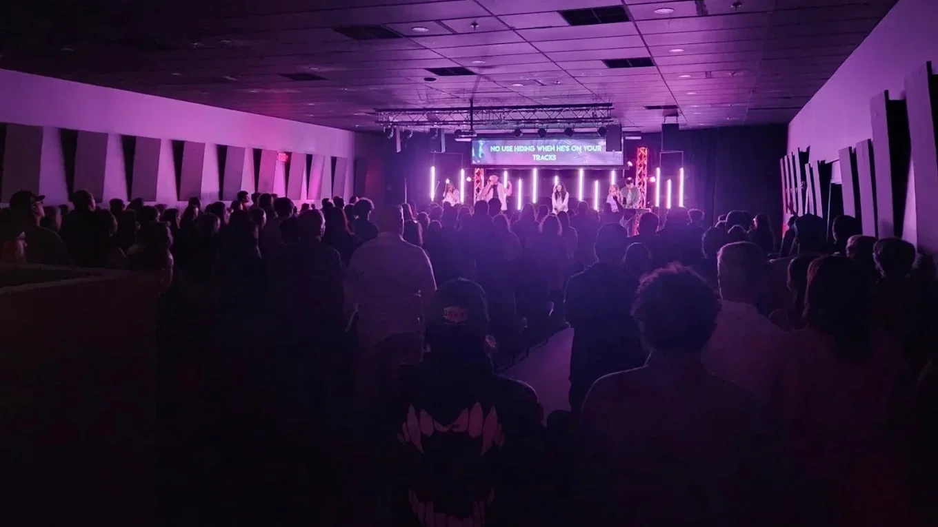 A large crowd in a dark church auditorium with purple lighting. There is a stage with band members and a large screen displaying lyrics or messages.