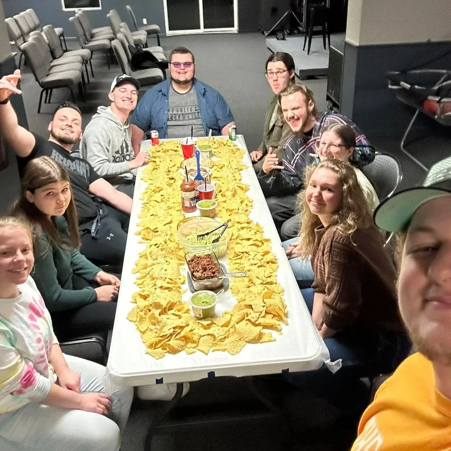 A group of people sitting around a long table covered with yellow chips, with various dips and condiments, celebrating at a life group.