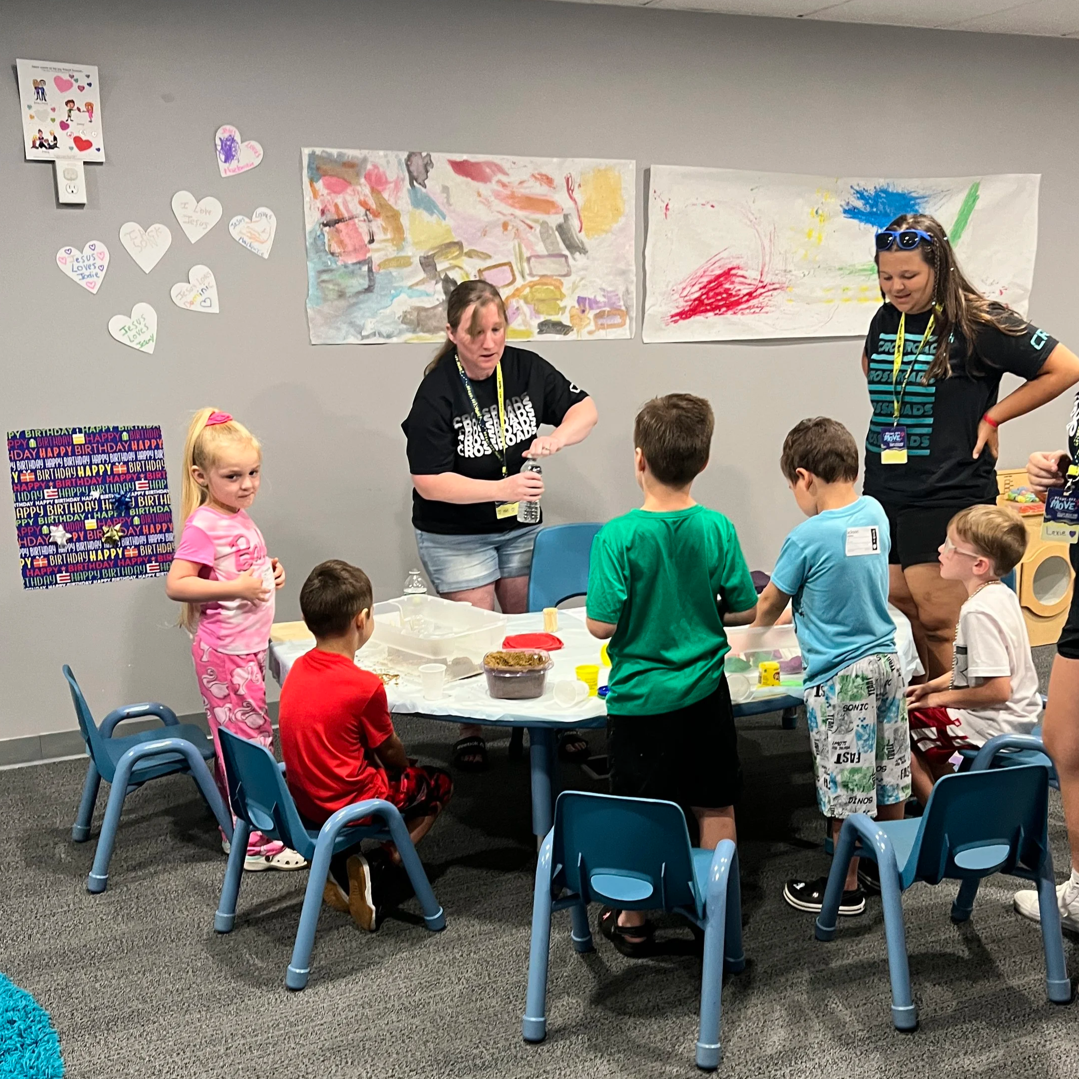 Children and adults gathered around a table with birthday decorations, eating cake and participating in activities in a room with colorful art on the wall.