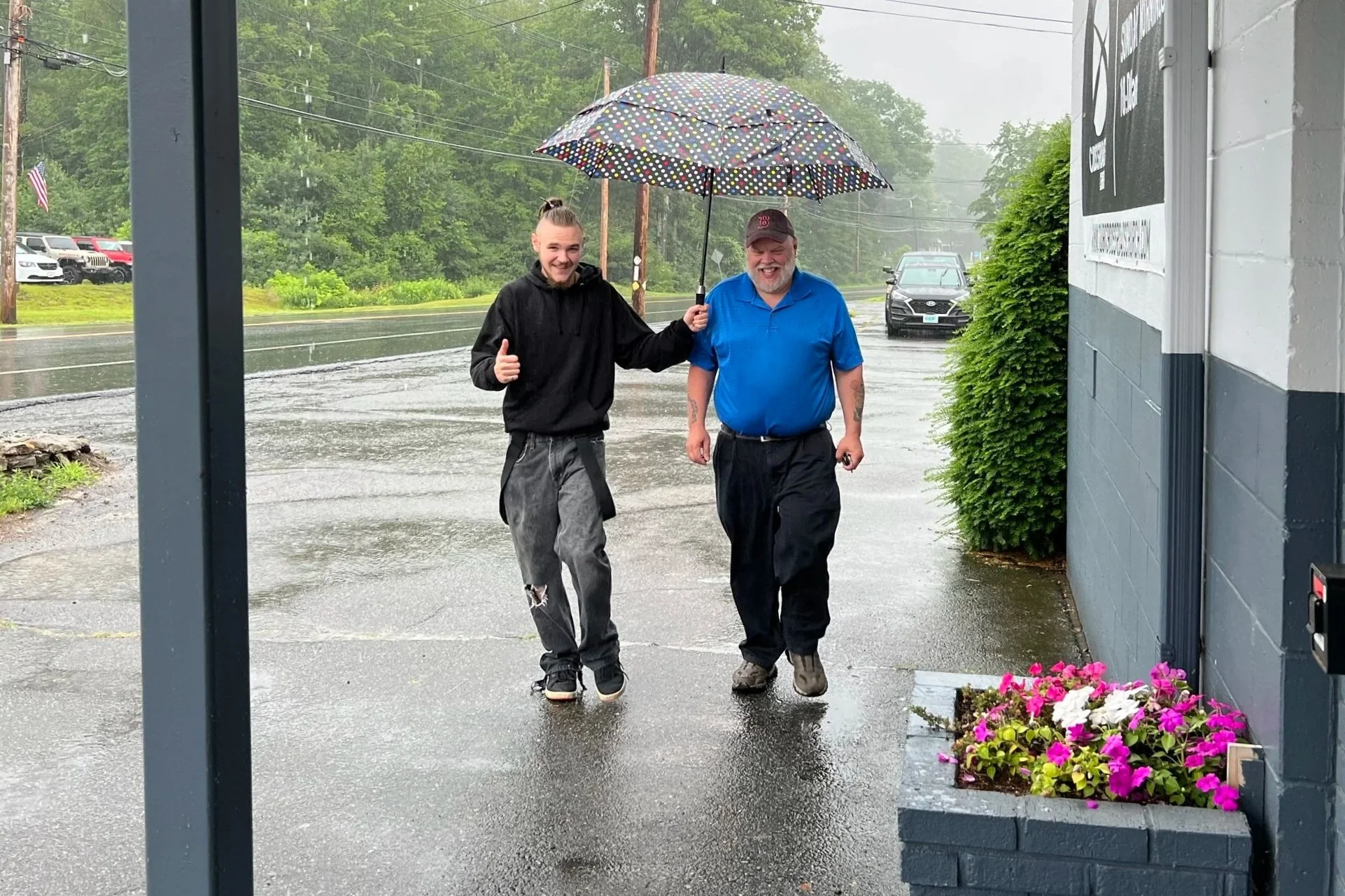 Two men walking in the rain under a polka dot umbrella, smiling and giving a thumbs up, with the rain hitting the pavement and cars parked along the street in the background.