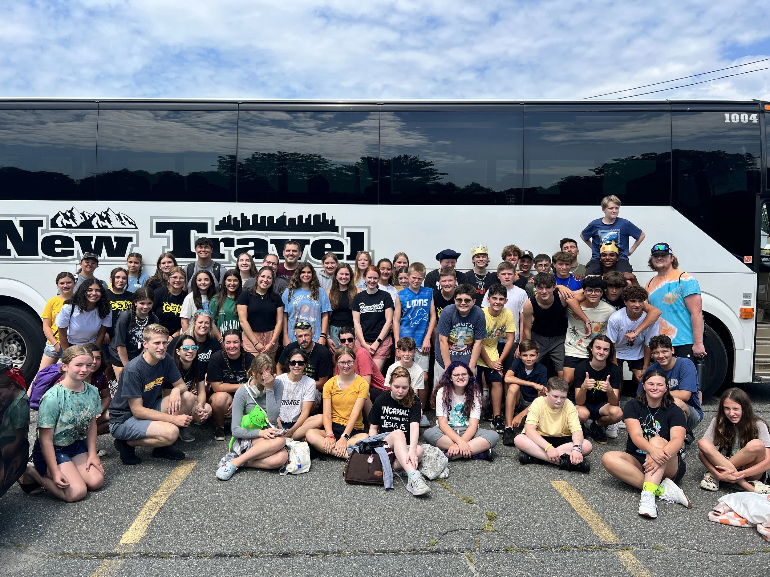A large group of children and adults with some wearing colorful shirts, gathered outside for a group photo in front of a bus. The bus is parked on a paved lot, with trees and a partly cloudy sky in the background.