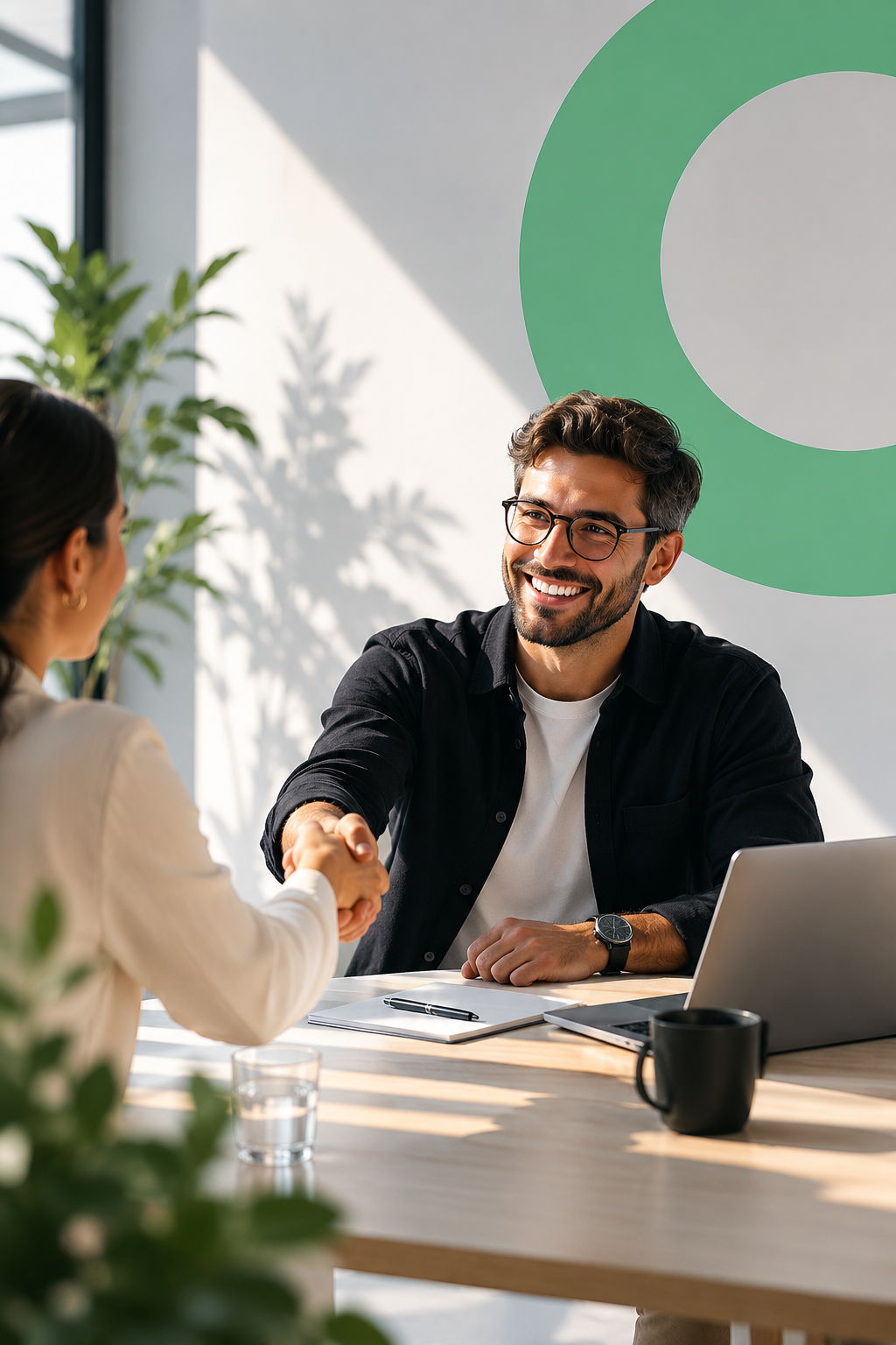 Man and woman shaking hands in an office setting, smiling, with a laptop, notebook, and coffee mug on the table, natural light, plants, and a graphic wall in the background.