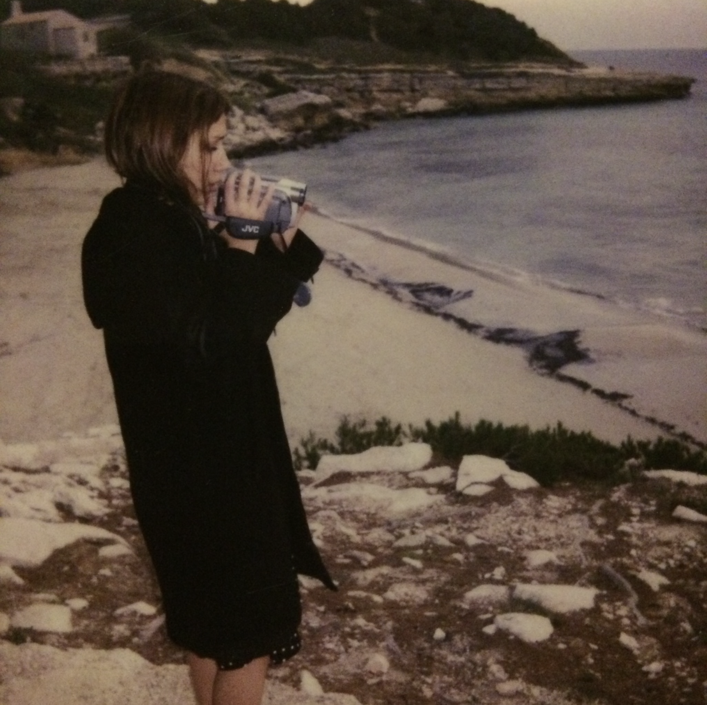 La cinéaste Amélie Derlon Cordina, à 17 ans, à la plage en soirée, portant un manteau noir, tenant une petite caméra pointée vers la mer, avec des rochers, du sable et des vagues en arrière-plan.