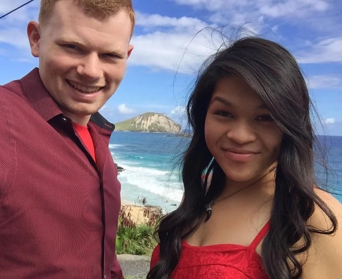 A smiling young man and woman taking a selfie outdoors near the ocean with a small island in the background, both wearing red tops.
