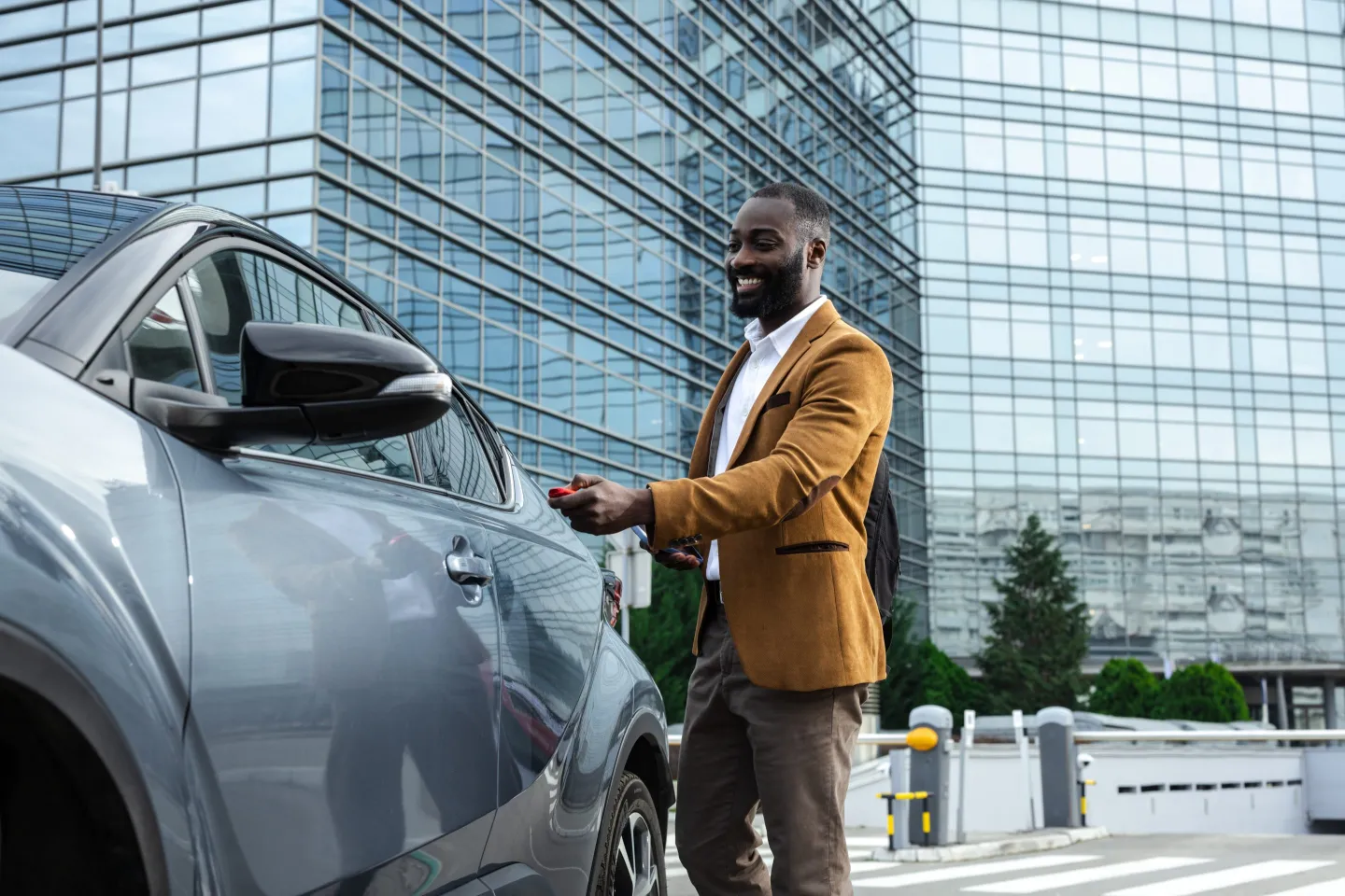A man in a brown blazer and white shirt standing outside in front of a modern glass office building, holding a smartphone and smiling.
