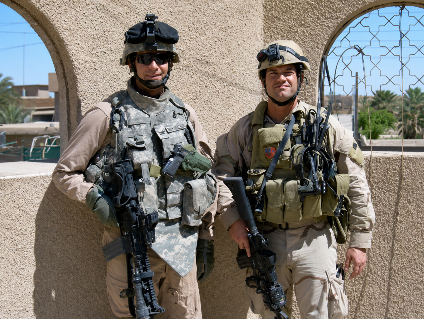 Two soldiers in uniform and tactical gear standing outdoors, with a beige wall and blue sky behind them.