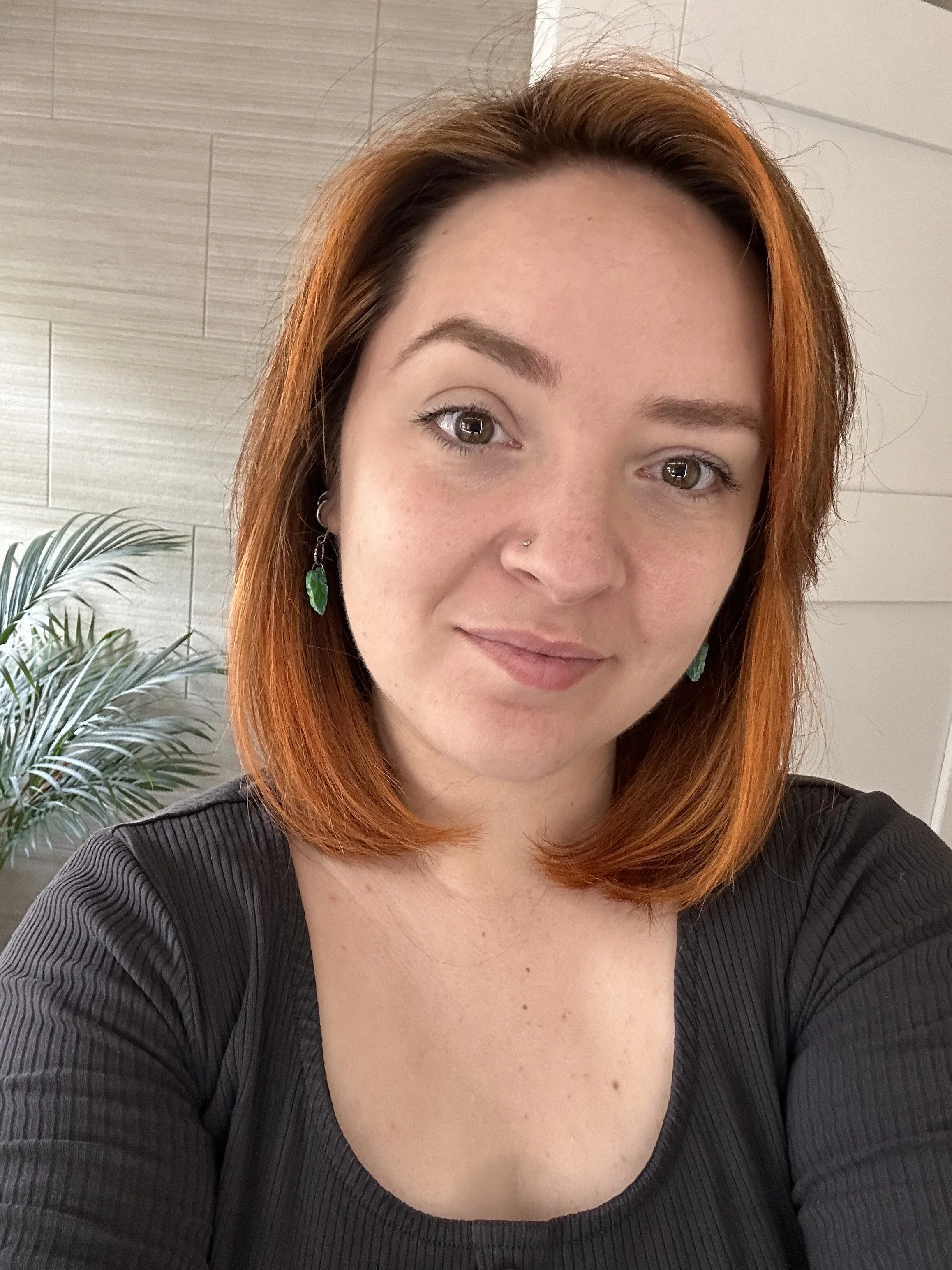 A woman with shoulder-length red hair, green leaf earrings, and a nose piercing is smiling slightly in a close-up selfie. She is wearing a black ribbed top, and part of a plant and a wooden wall are visible in the background.
