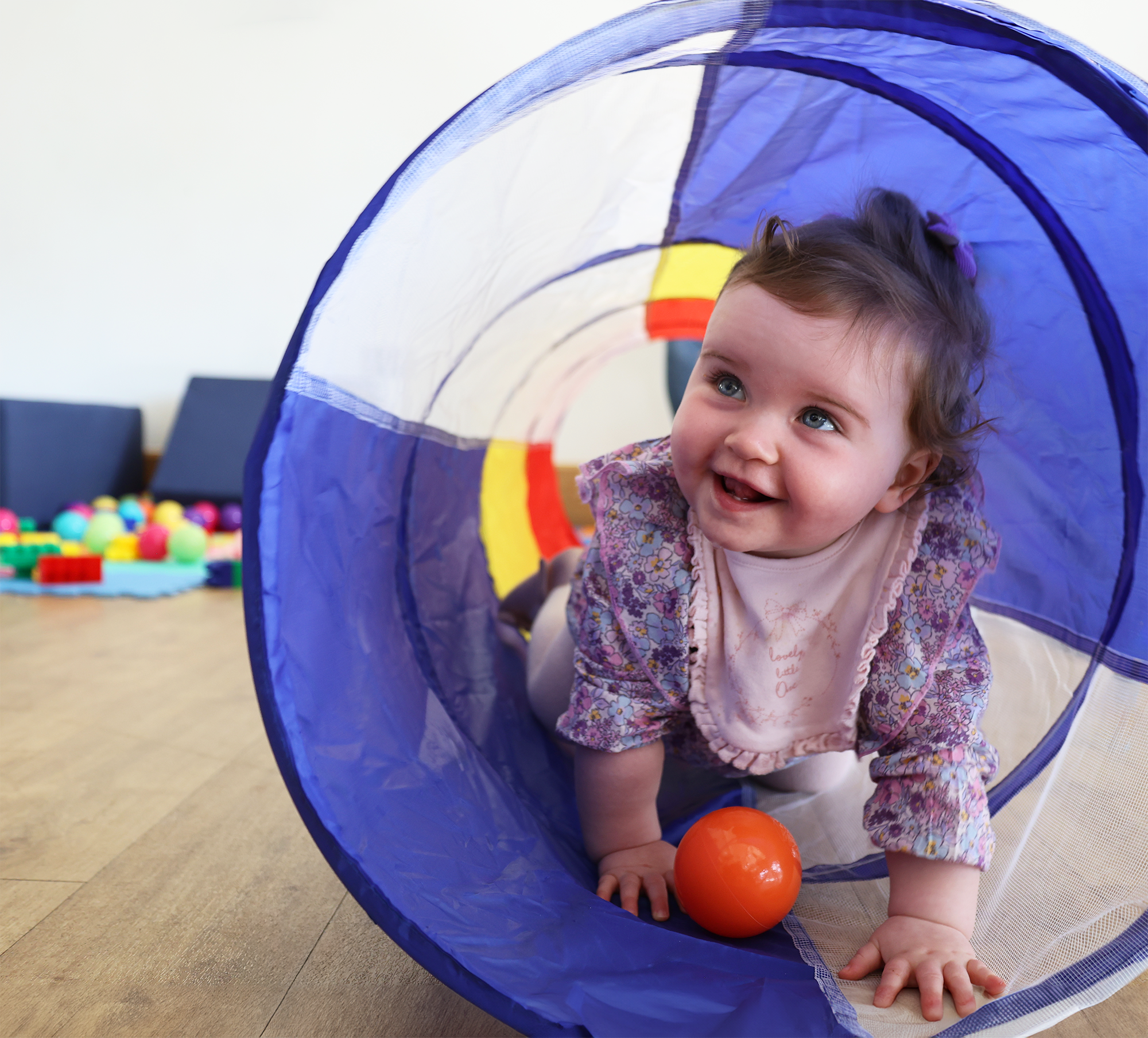 A young girl with brown hair and blue eyes playing inside a colorful play tunnel with orange and yellow balls scattered around. She is wearing a floral-patterned shirt and a white bib, crawling on a wooden floor in an indoor setting.