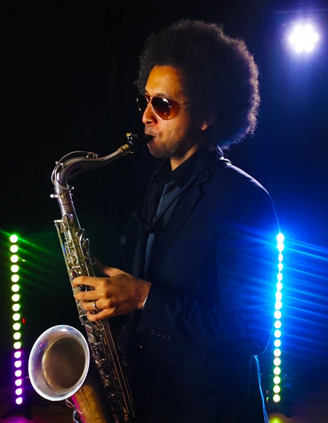 A man with an afro hairstyle wearing sunglasses and a suit, playing a saxophone on stage with colorful lights.