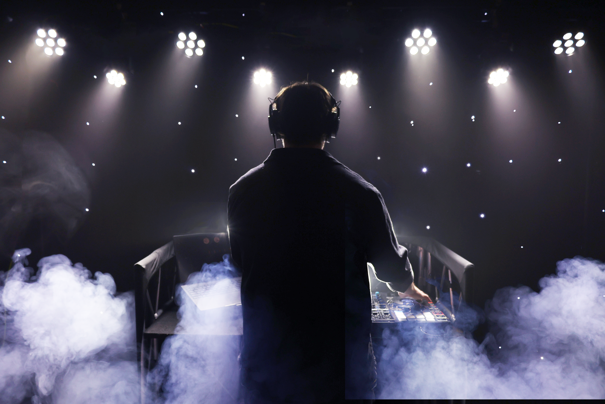 Silhouette of a DJ wearing headphones playing music at an event with bright stage lights and smoke effects.