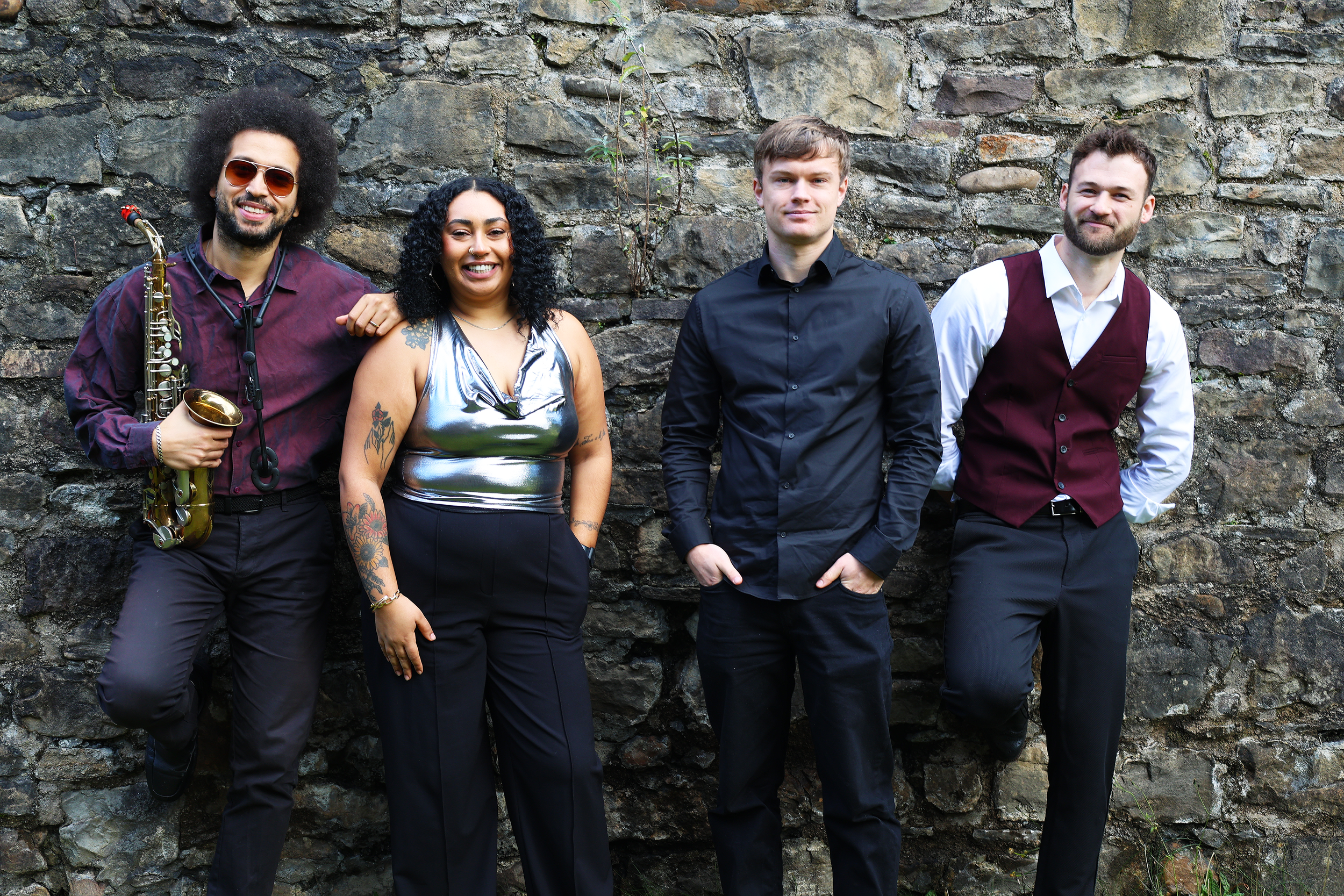 Group of four diverse musicians standing against a stone wall, smiling, with one holding a saxophone.