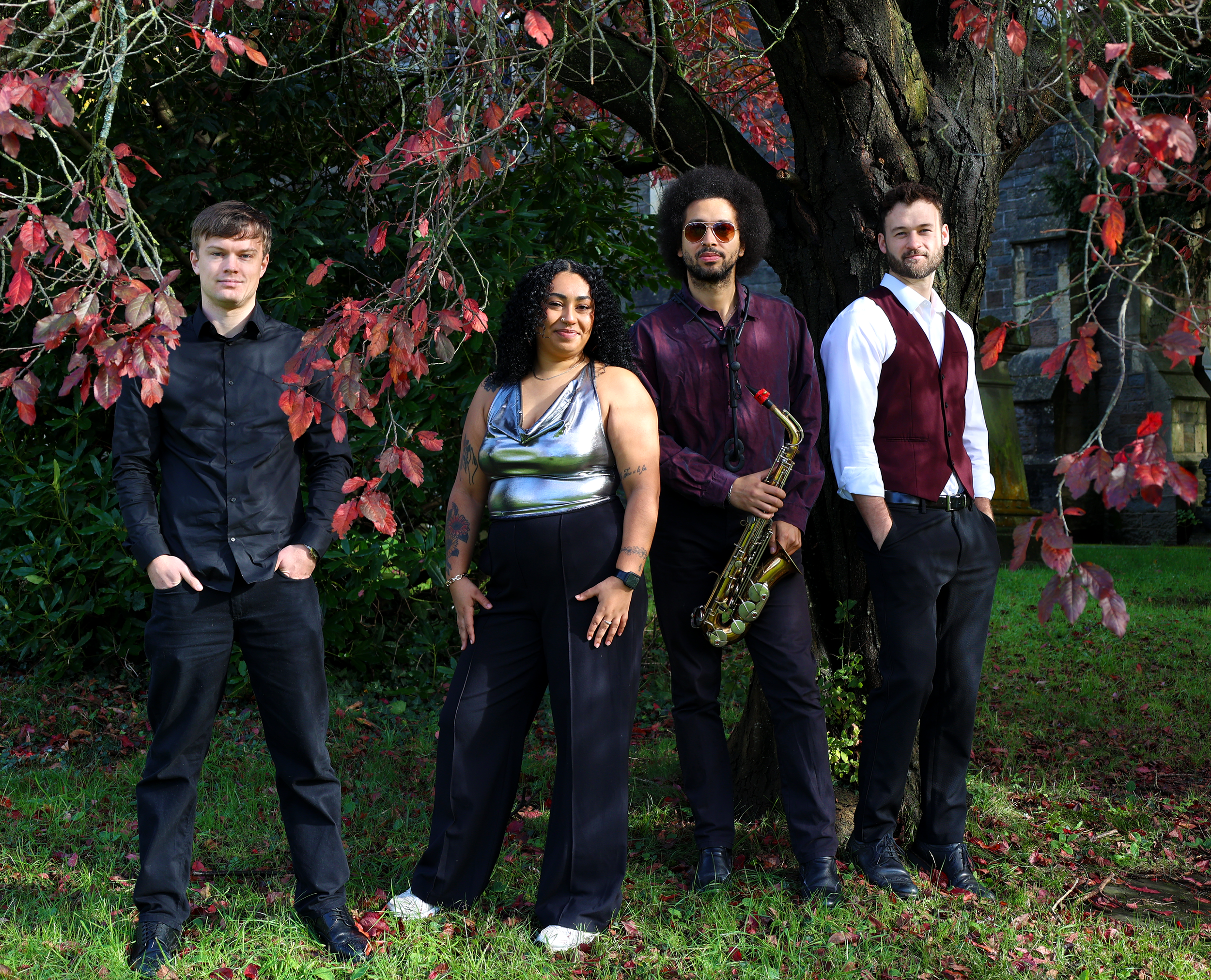 Four musicians standing outdoors in front of a large tree with red and green leaves. The group includes three men and one woman, with one man holding a saxophone. All are dressed semi-formally, and the weather appears mild.