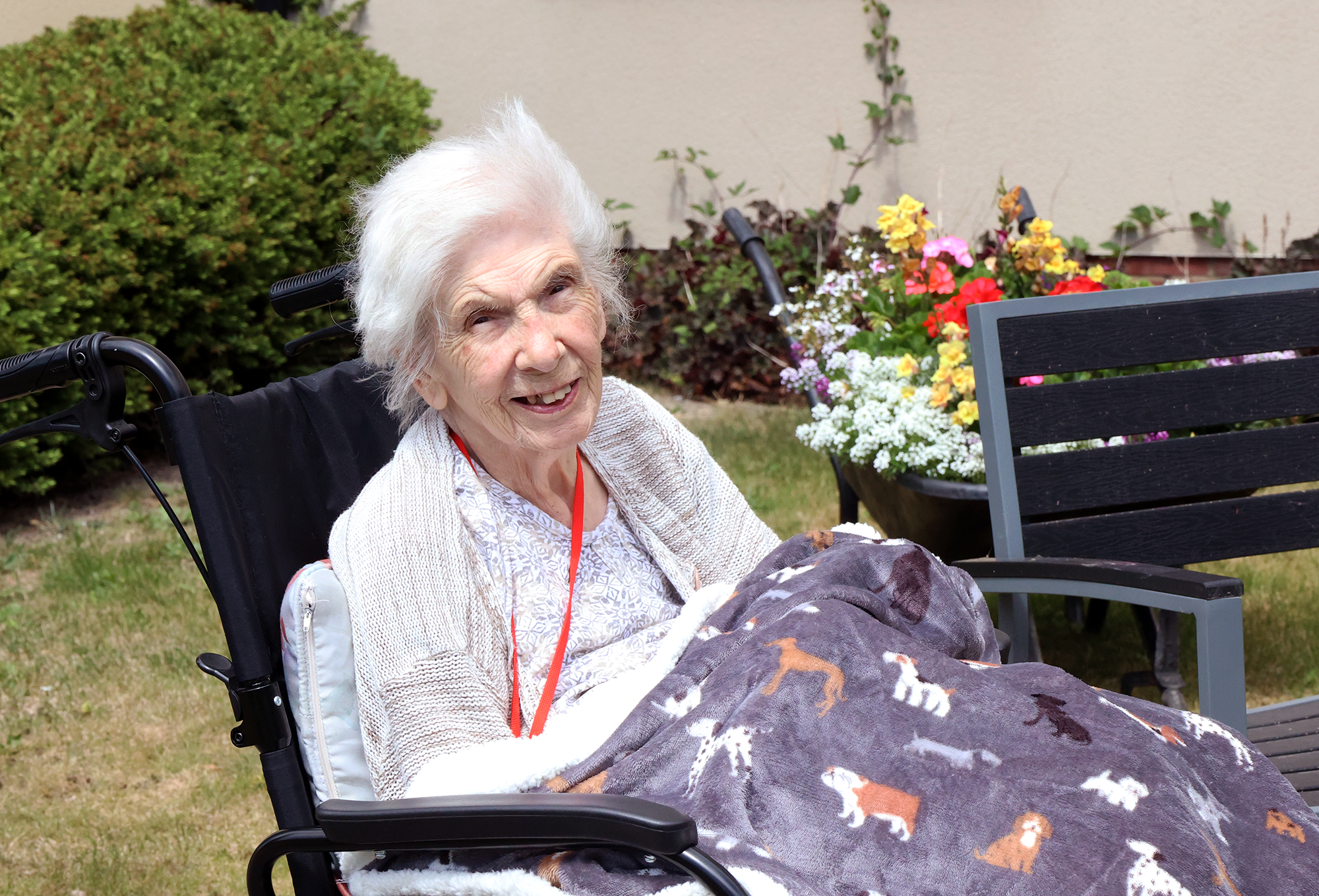 An elderly woman with white hair smiling while sitting outside in a wheelchair, wrapped in a blanket, with colorful flowers and bushes in the background.