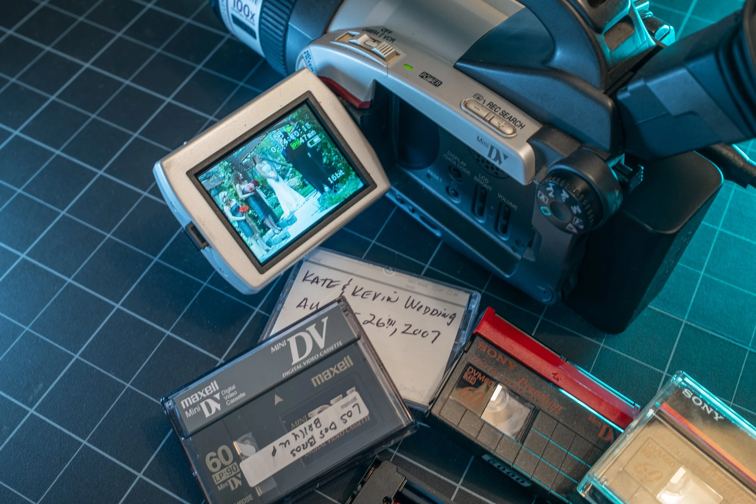 A video camera with its flip-out screen showing a wedding scene with people standing outdoors under trees. The camera is placed on a black grid-patterned surface, surrounded by various VHS tape cases.