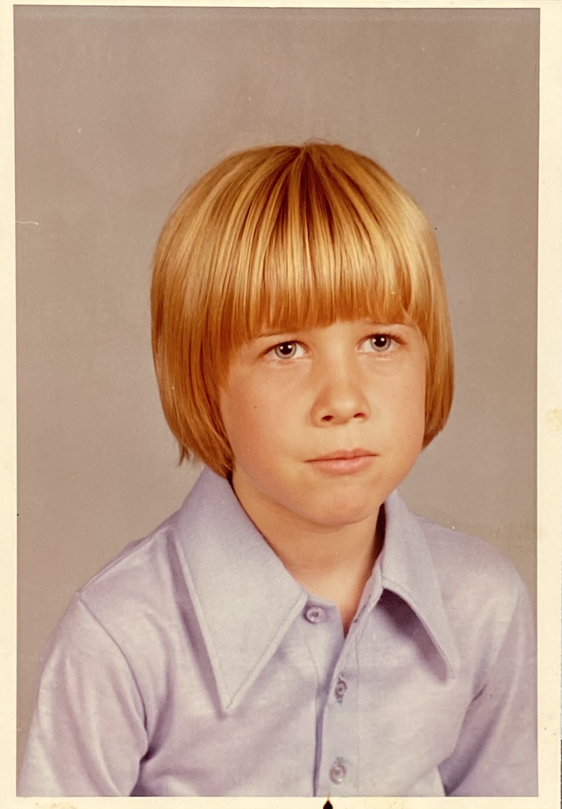 A young boy with straight red hair, wearing a light purple collared shirt, looking directly at the camera.