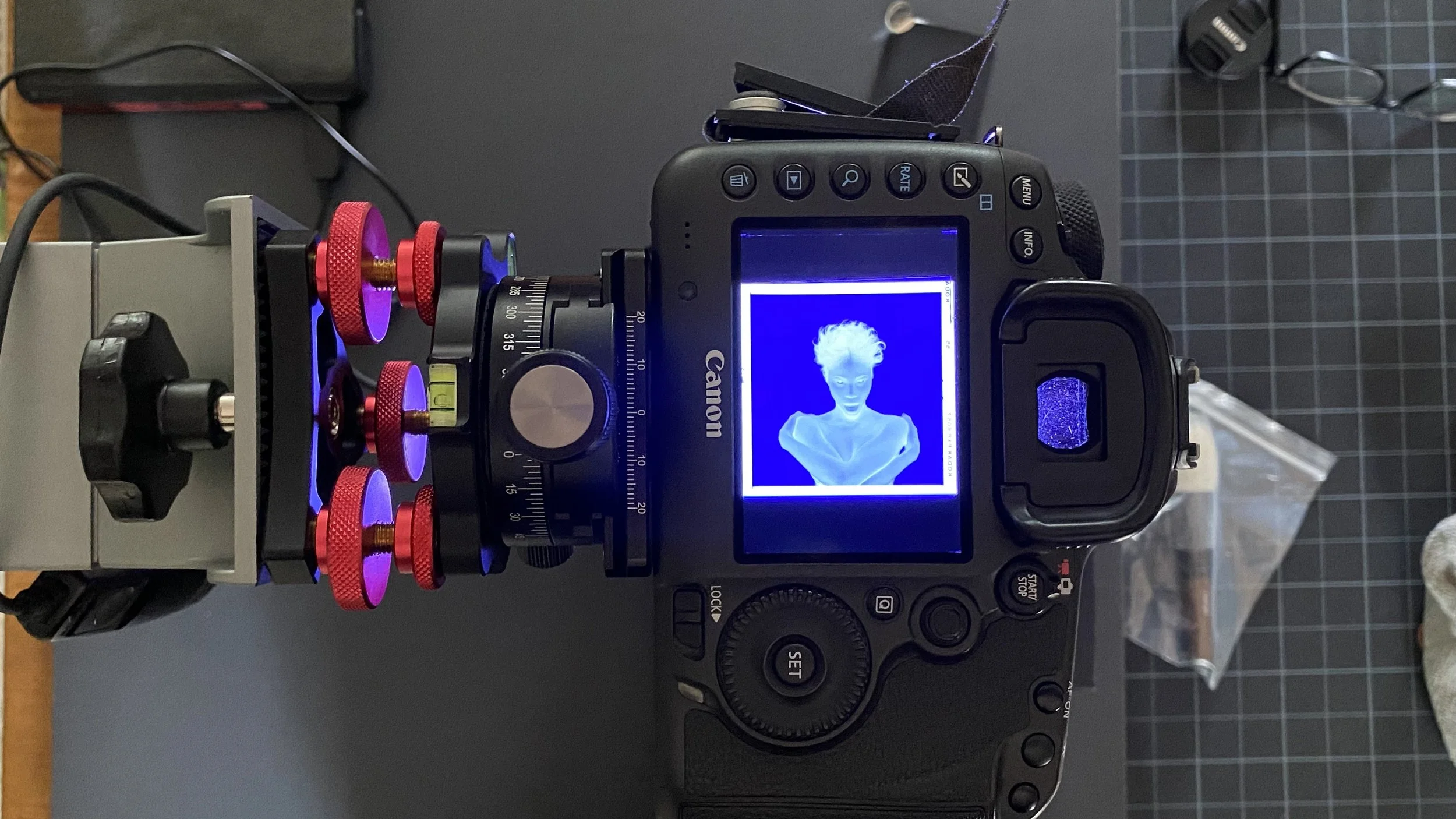A Canon DSLR camera with a digital screen displaying a photo of a woman with short, wild hair and an open-mouthed expression. The camera is mounted on a fixture with multiple red and purple components, and there are various objects and cables on the desk nearby.