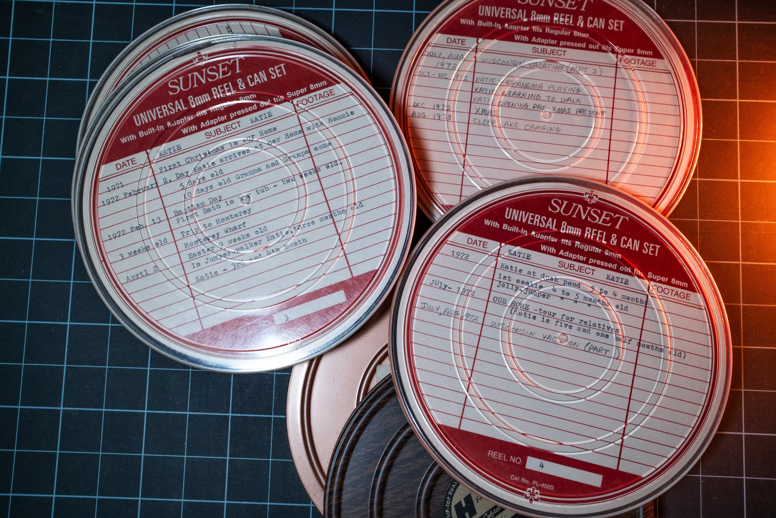 Close-up view of three round red and clear plastic reels with handwritten labels on top, placed on top of a blue cutting mat with grid lines. The reels are for 8mm film, with writing indicating subjects and dates.