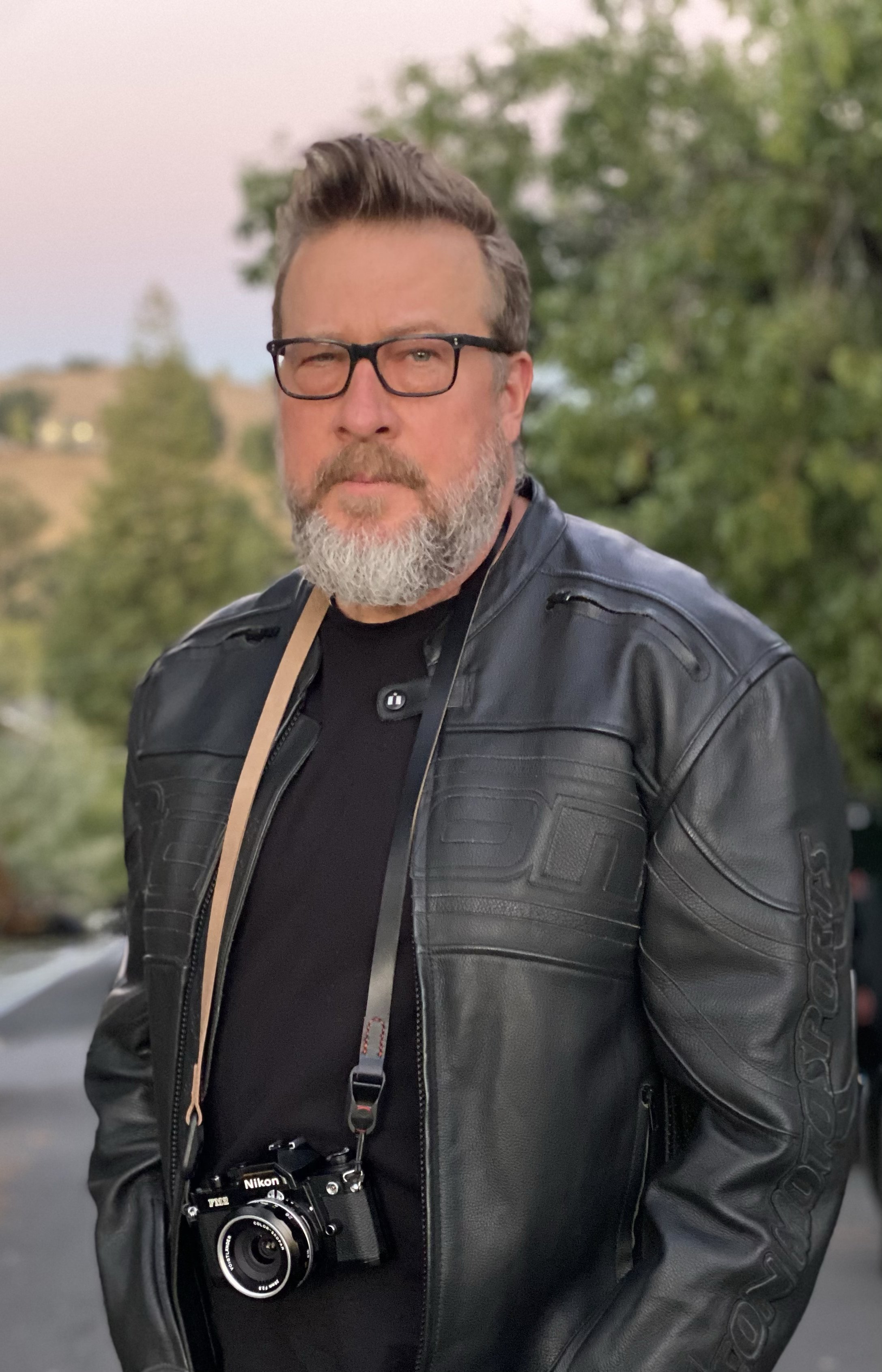 A man with a beard and glasses wearing a black leather jacket, standing outdoors with trees and a clear sky in the background, holding a Nikon camera around his neck.