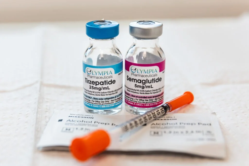 Two vials labeled Tirzepatide 25mg/mL and Semaglutide 5mg/mL, a syringe with an orange cap, and alcohol prep pads on a white surface.