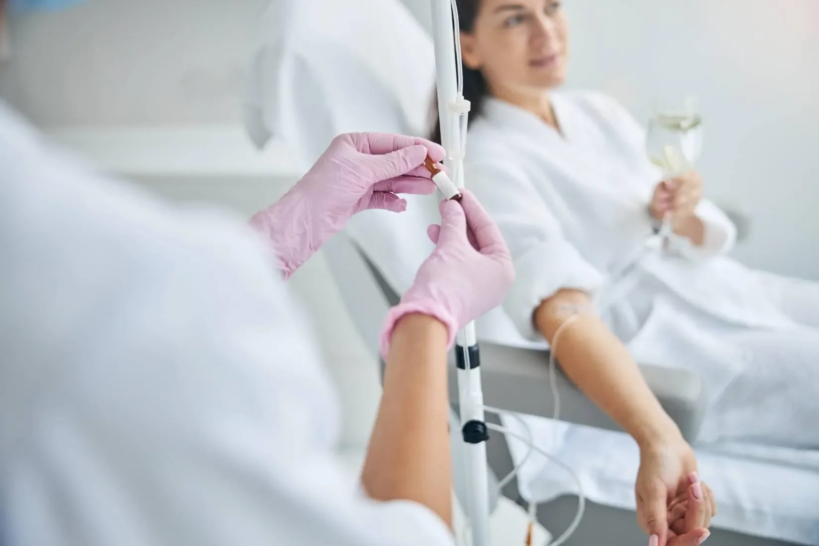 Close-up of a nurse or healthcare worker with pink gloves in a hospital, preparing an injection, with a woman patient in the background who is receiving IV fluids and holding a glass of water.