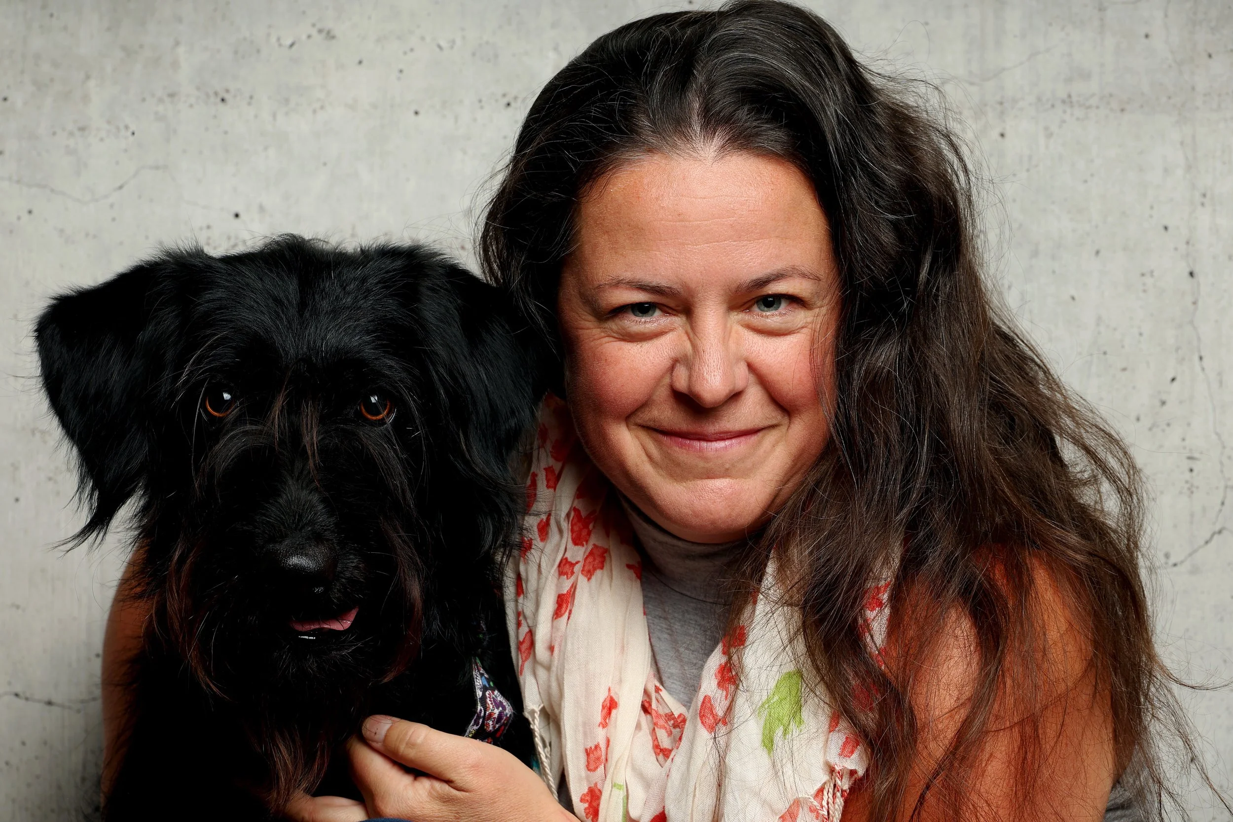 Une femme avec des cheveux bruns longs et une peau claire, souriante, tenant un chien noir avec des yeux marron et portant un collier en tissu, devant un mur en béton.