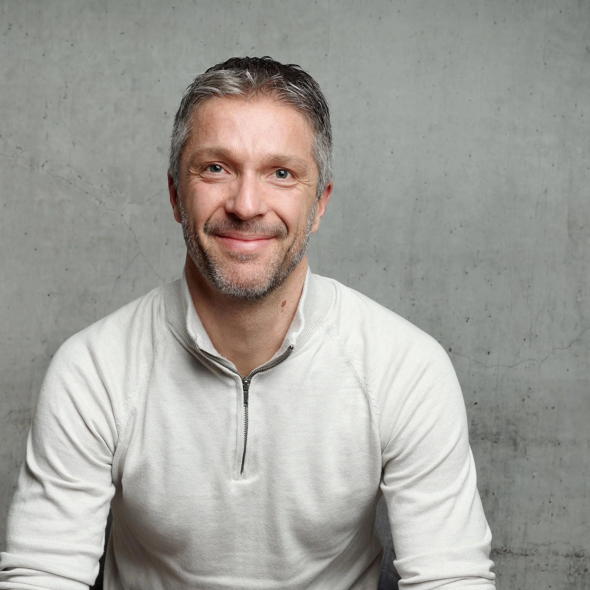 Un homme mature avec des cheveux gris, portant un pull blanc avec une fermeture éclair, souriant et assis devant un mur en béton gris.