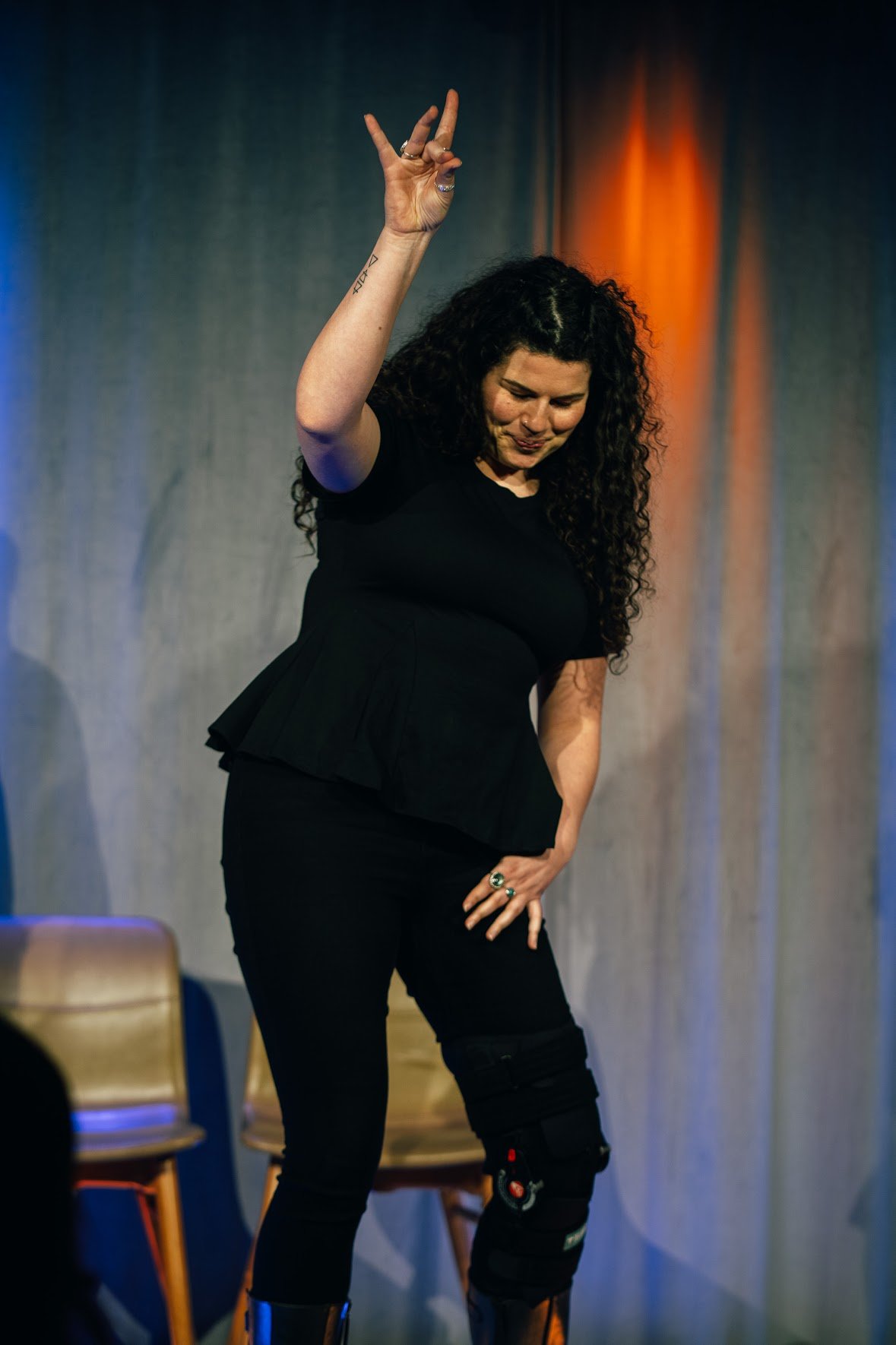 A woman with curly dark hair performing on stage, wearing black clothing and knee pads, with her right hand raised making a peace sign and her head bowed.