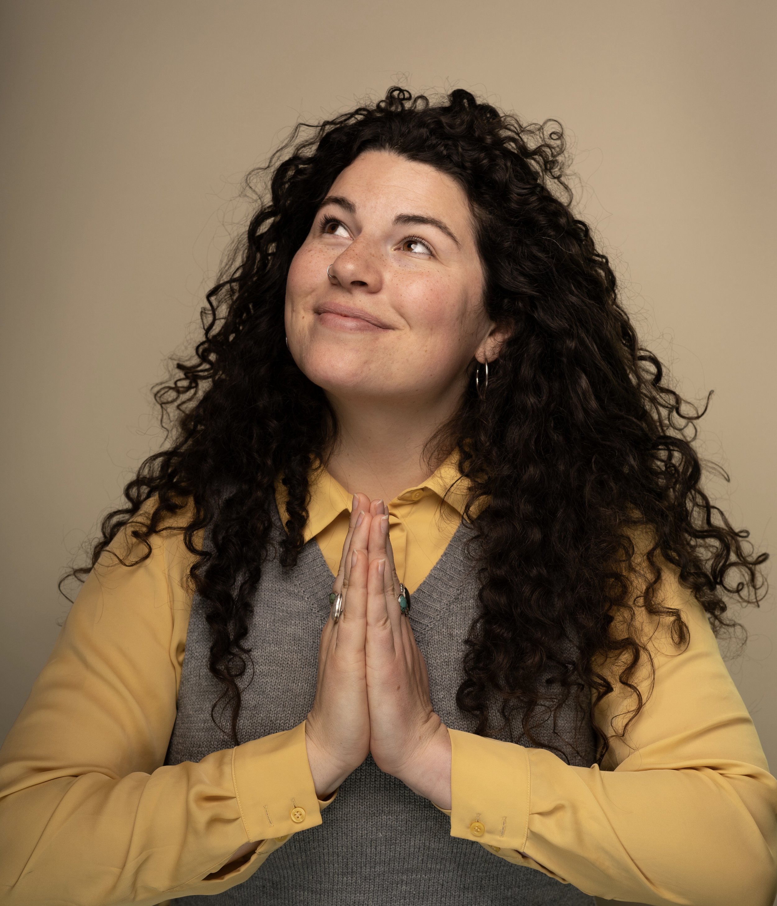 A woman with curly dark hair, wearing a yellow shirt and a gray vest, holding her hands in a prayer position near her chest, looking up with a peaceful expression.