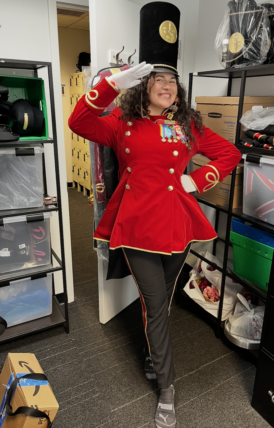 Young woman dressed in a red band uniform with medals, saluting and smiling in a storage room with shelves and boxes.
