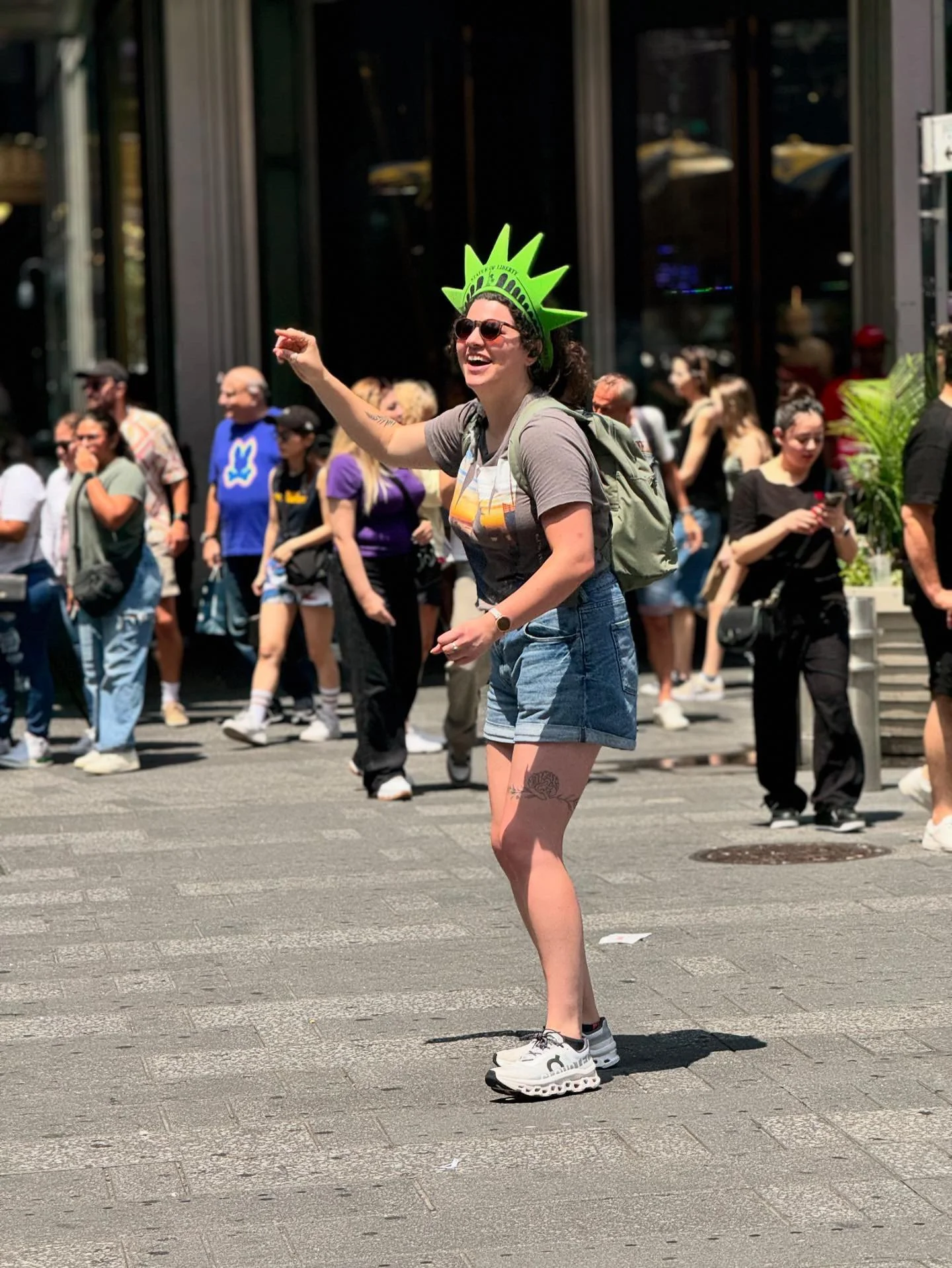 A woman with curly hair wearing sunglasses, a gray t-shirt, denim shorts, and white sneakers is standing in the street, smiling and waving in front of a crowd of people. She is wearing a green crown and carrying a backpack.