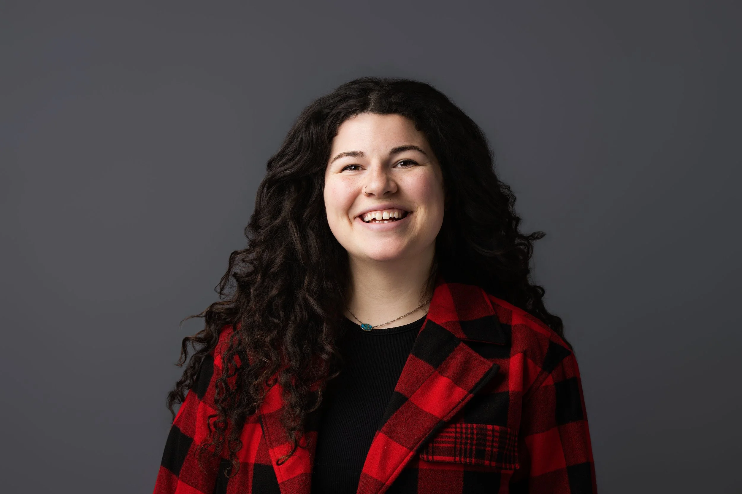 A woman with long curly dark hair, smiling, wearing a black top, red and black plaid jacket, and a small necklace, against a gray background.