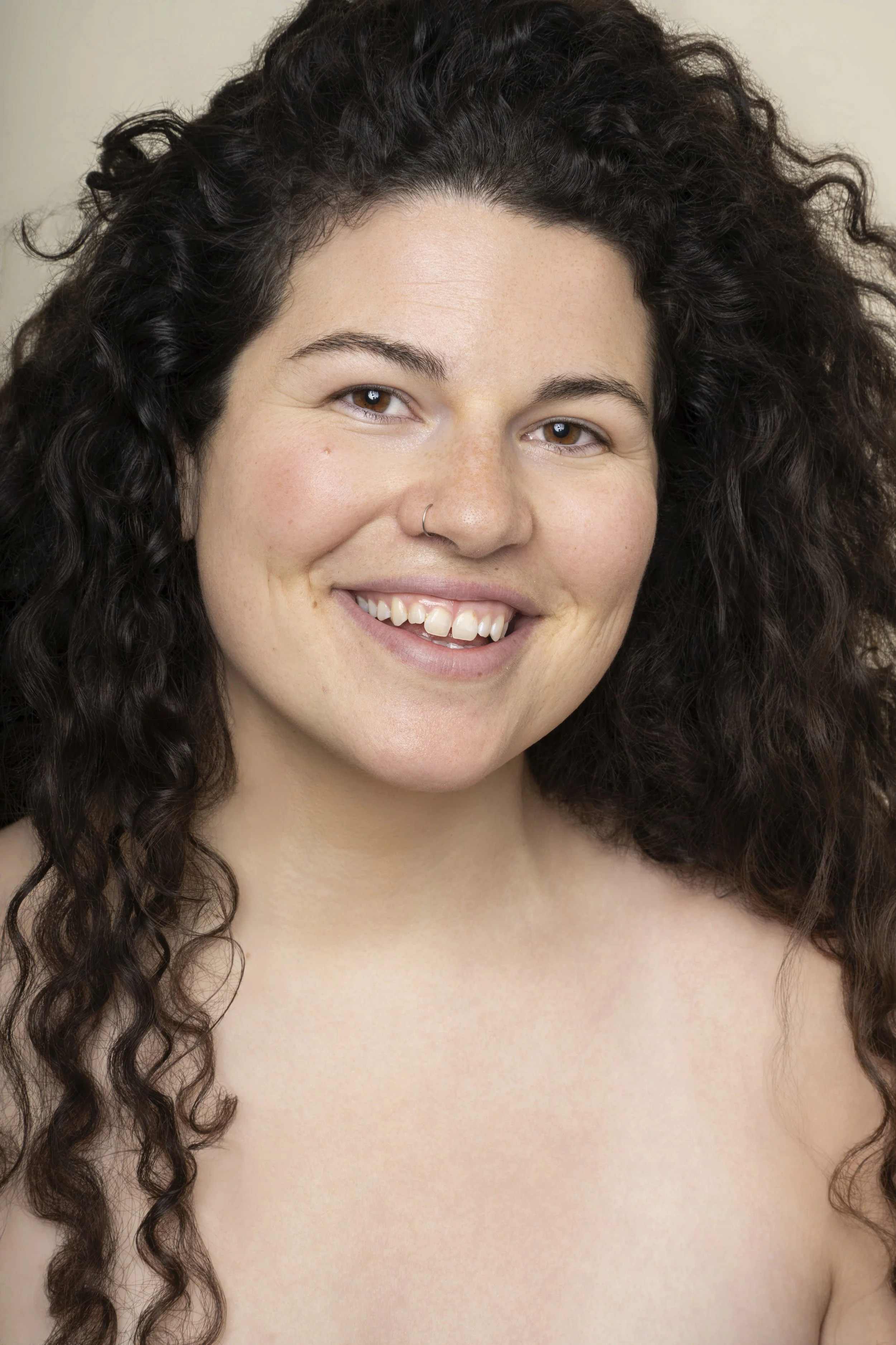 Close-up of a smiling woman with dark curly hair, light skin, and a small nose ring, topless with bare shoulders visible.