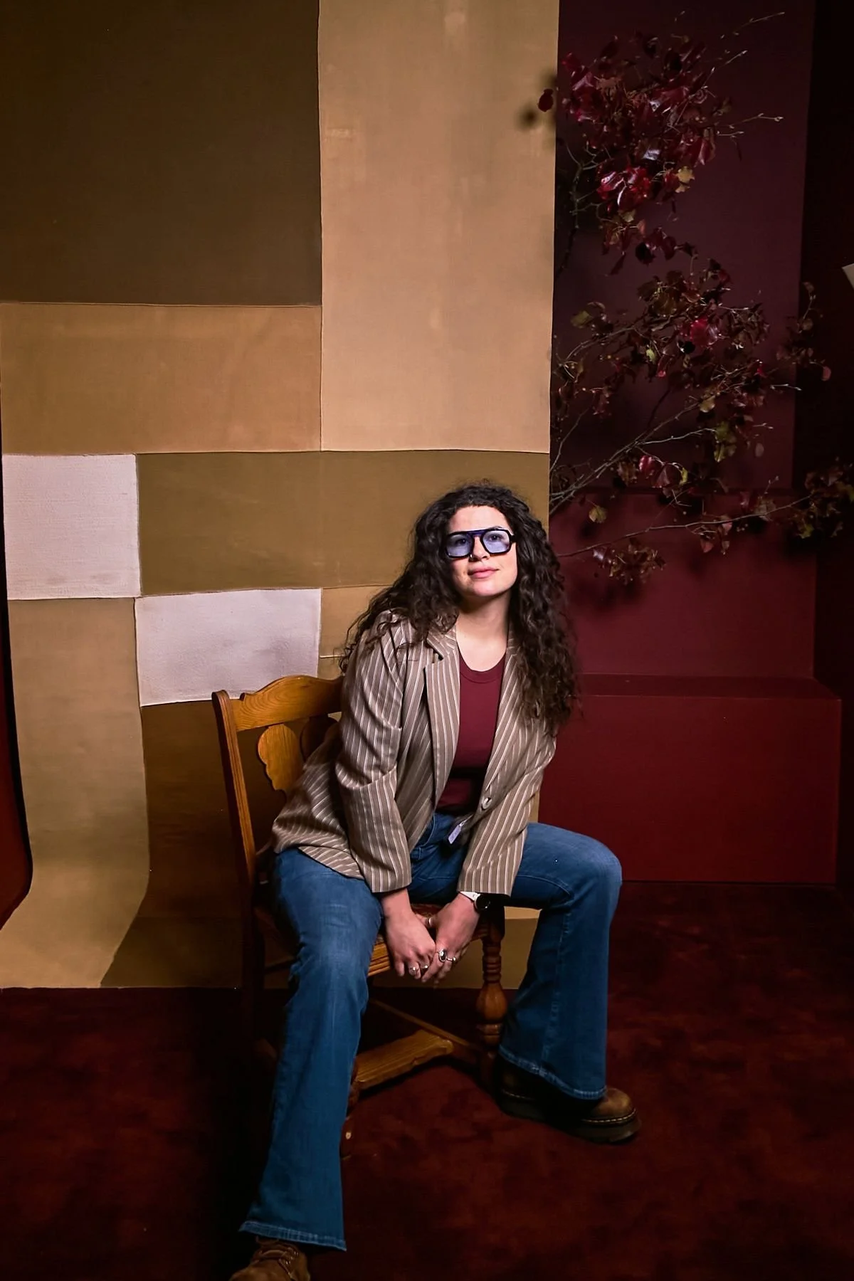 A woman with curly dark hair, wearing sunglasses, a striped blazer, a maroon top, and jeans, sitting on a wooden chair in front of a two-toned wall with a plant on the right side.