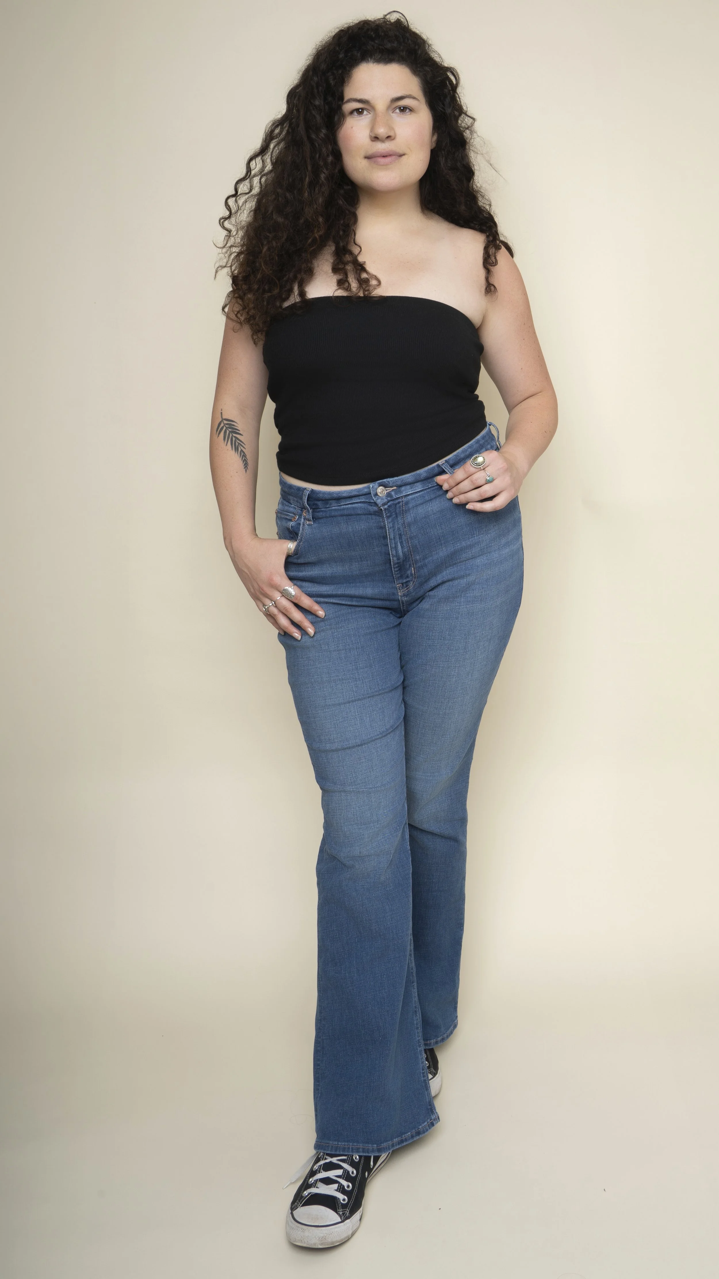 A woman with curly dark hair, wearing a strapless black top, blue jeans, black sneakers with white soles, and rings on her fingers, standing against a plain beige background.