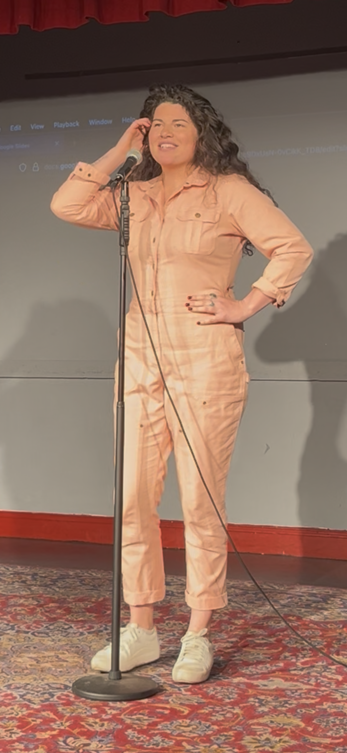 Woman with curly hair in peach-colored jumpsuit standing with hand on hip behind microphone on stage.