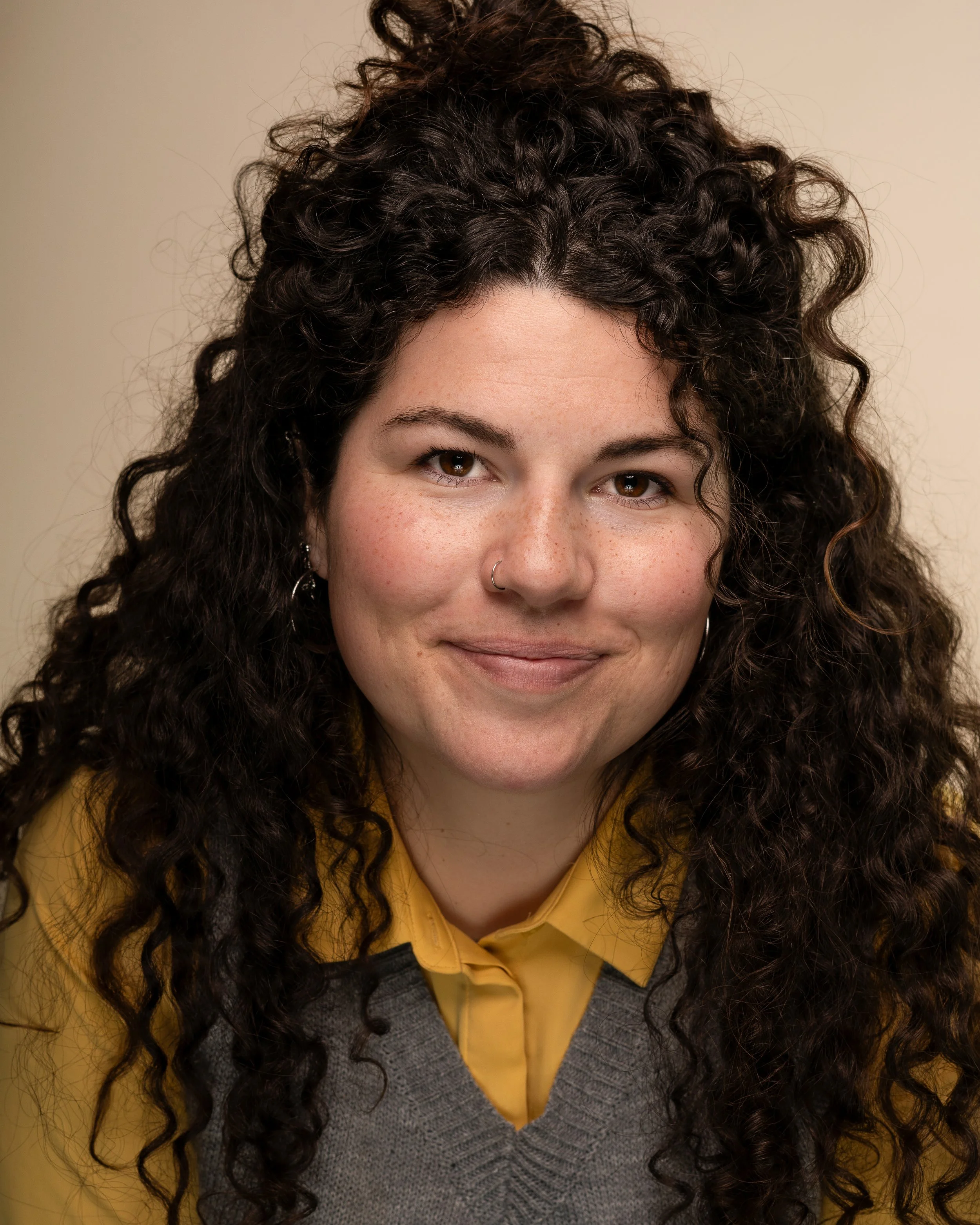 Close-up of a woman with long curly dark hair, wearing a yellow shirt and gray sweater, smiling gently.