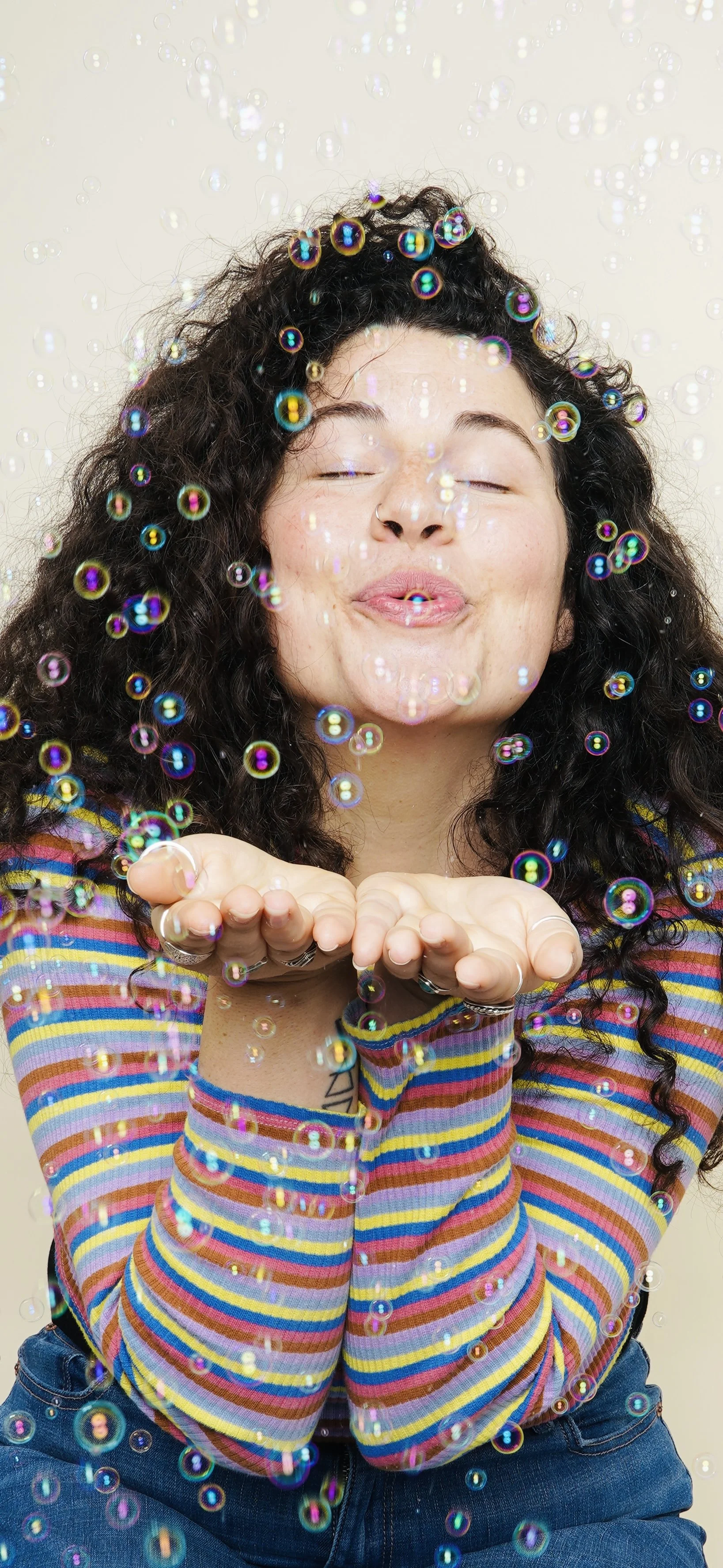 Woman with curly hair blowing bubbles and smiling with eyes closed, wearing a colorful striped shirt and rings on her fingers.